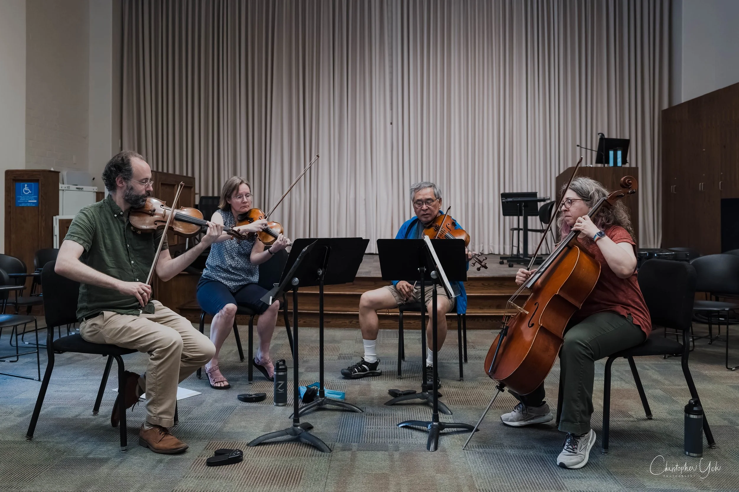 Group of five musicians practicing with violins and a cello on a stage with music stands and curtains in the background.