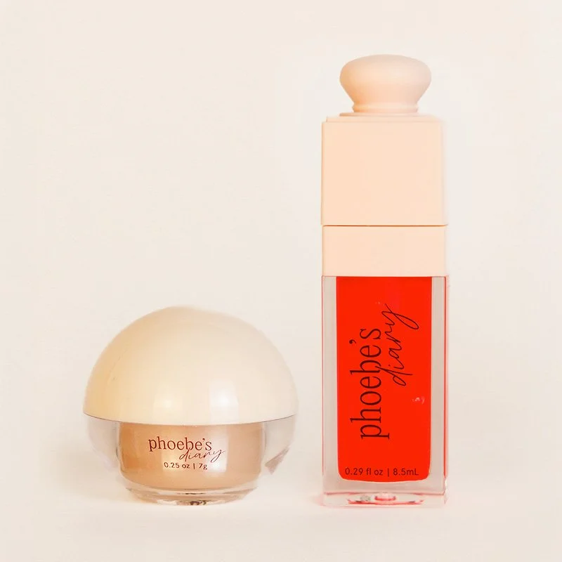 Two jars of Phoebe's Diary cosmetic product, one small round container with a beige lid and another taller rectangular container with a blush pink cap and a red label.