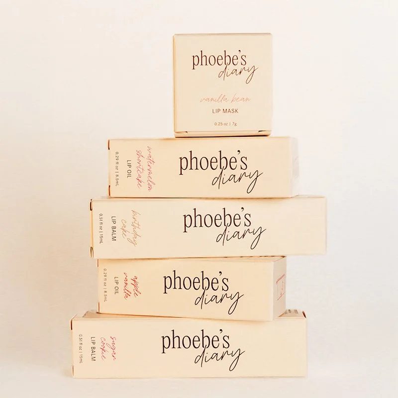 Stacked boxes of Phoebe's Diary beauty products, including vanilla bean lip mask, lavender lip balm, rose lip oil, and specialty lip balm.