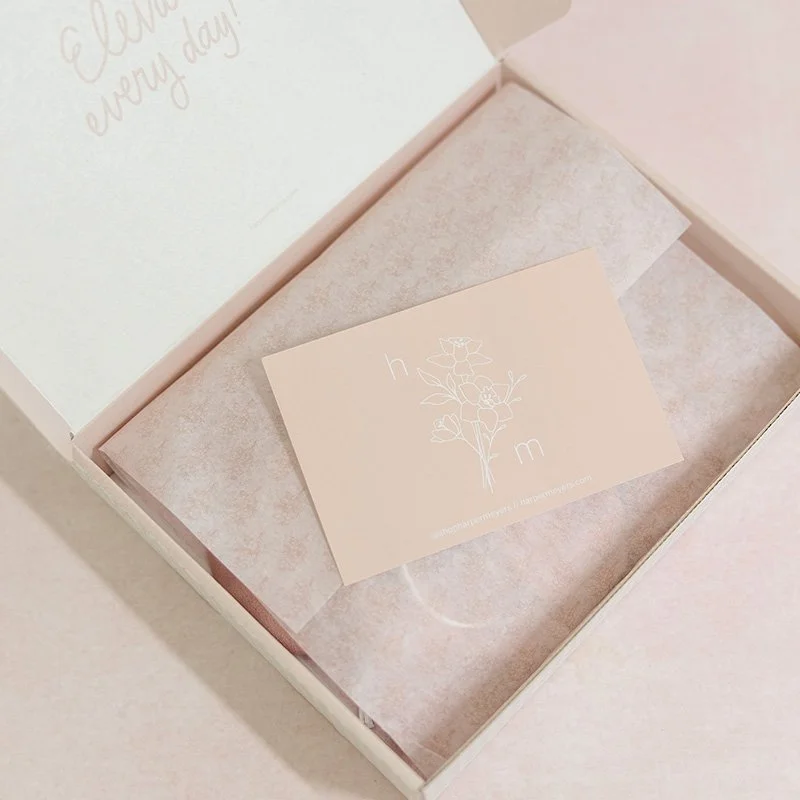 Open pink gift box with tissue paper and a small card inside, featuring a white floral illustration and the letters 'h' and 'm'.