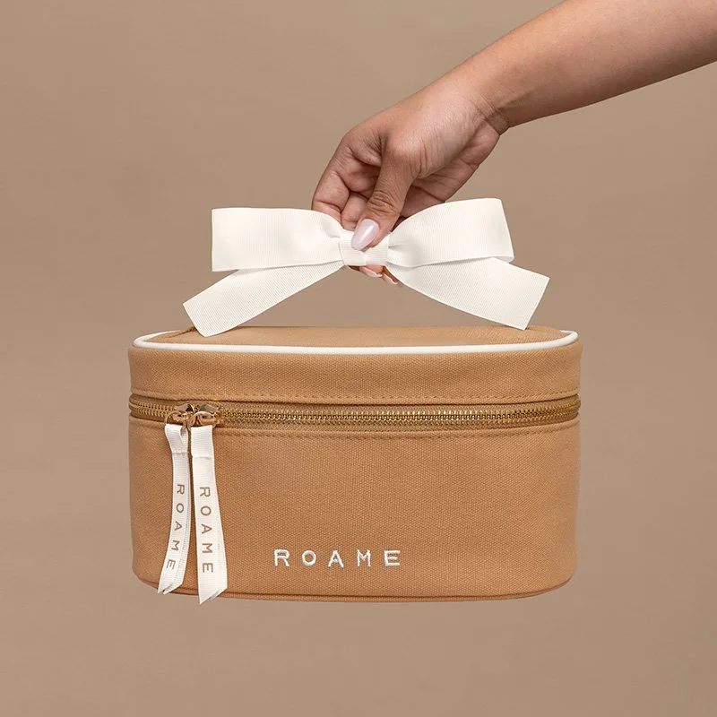 A person's hand holding a white ribbon tied in a bow on top of a beige toiletry bag with the brand name 'ROAME' embroidered on it, set against a plain background.