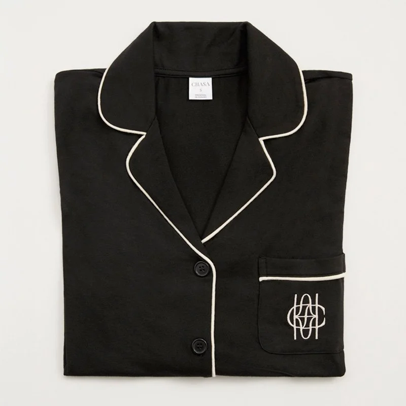 Black pajamas with white piping, folded neatly with a chest pocket embroidered with a white monogram, lying flat on a white background.