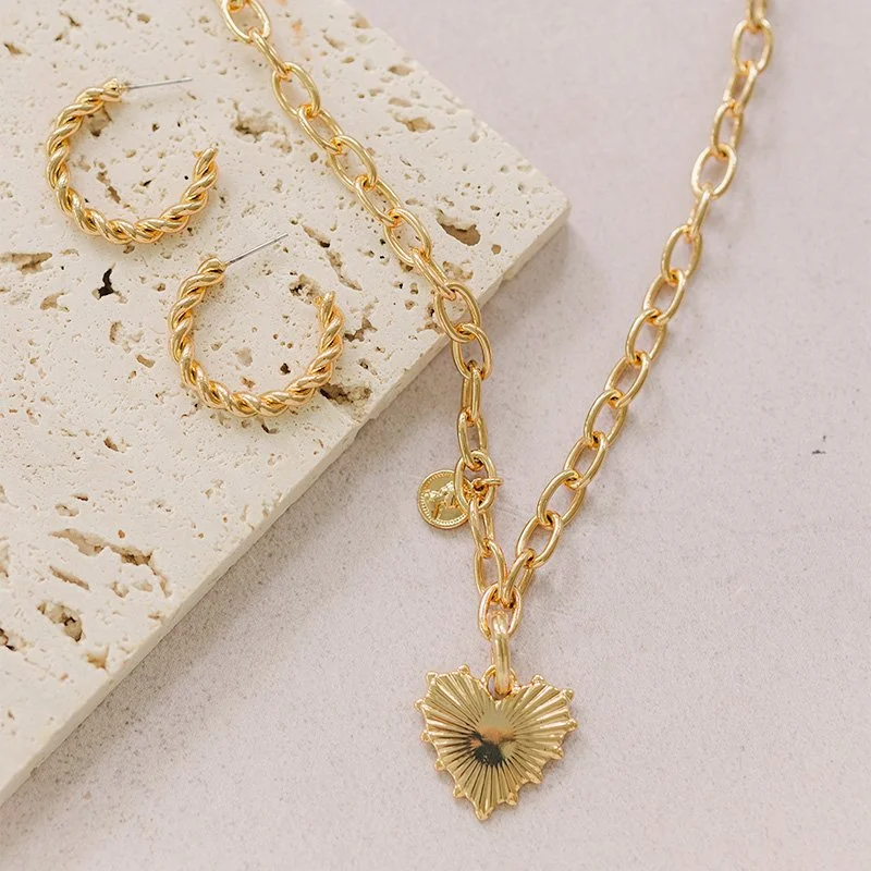 Gold jewelry set consisting of a chain necklace with a heart-shaped pendant, a pair of matching hoop earrings, all displayed on a white textured surface.