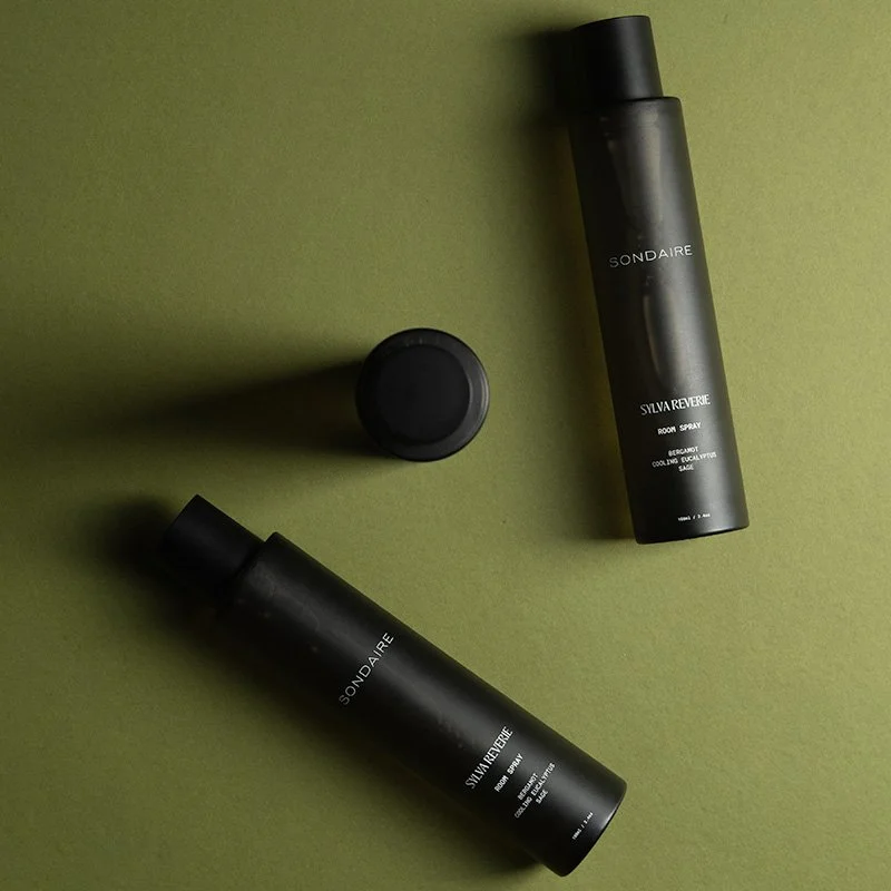 Two black bottles of SONDÀIRE room spray, one lying down and one standing, with a black cap placed on the green surface.