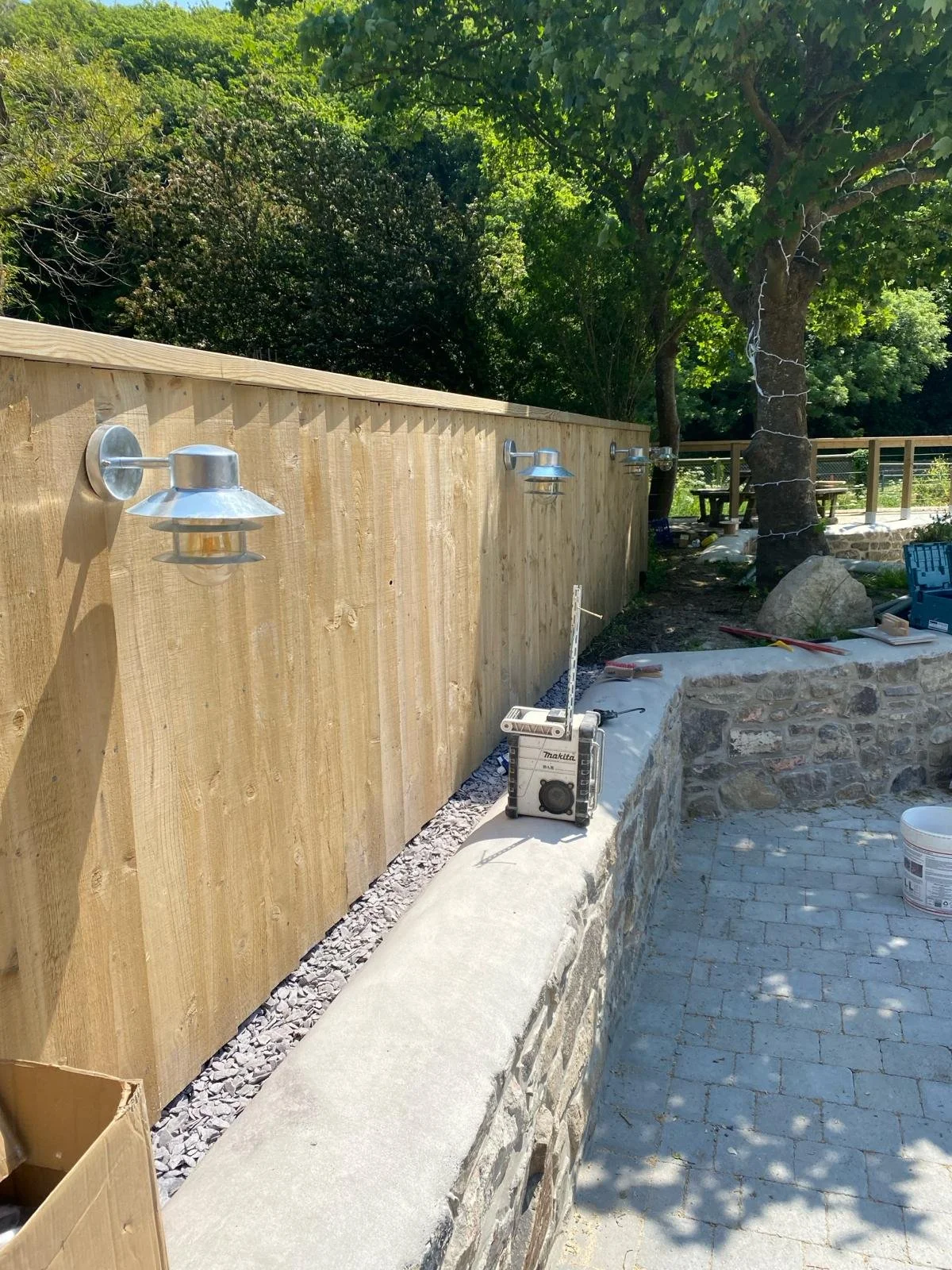 Construction site with a newly installed wooden fence, outdoor lighting fixtures, and stonework along a brick pathway, surrounded by trees.