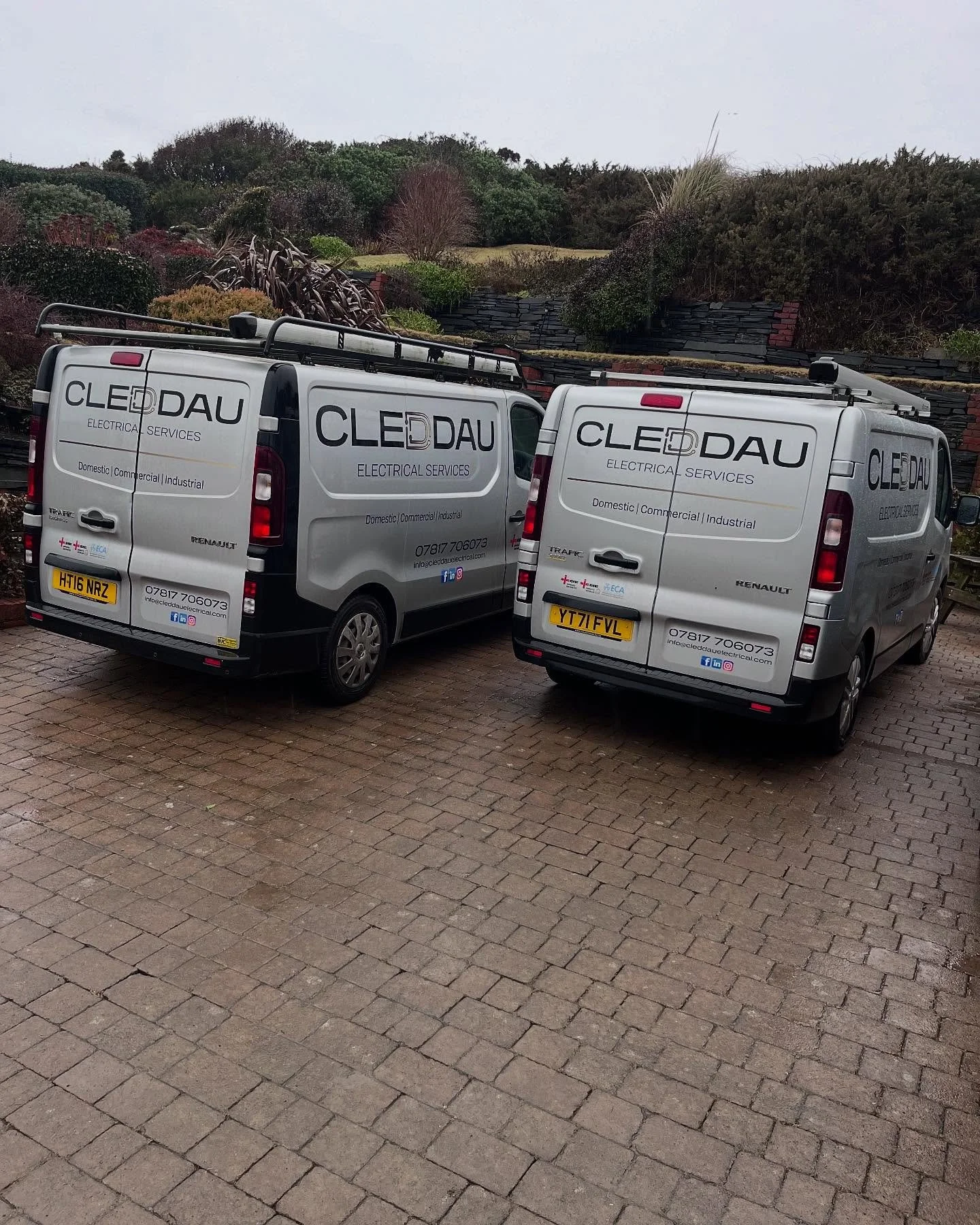 Two company vans parked on a paved area with bushes and trees in the background.