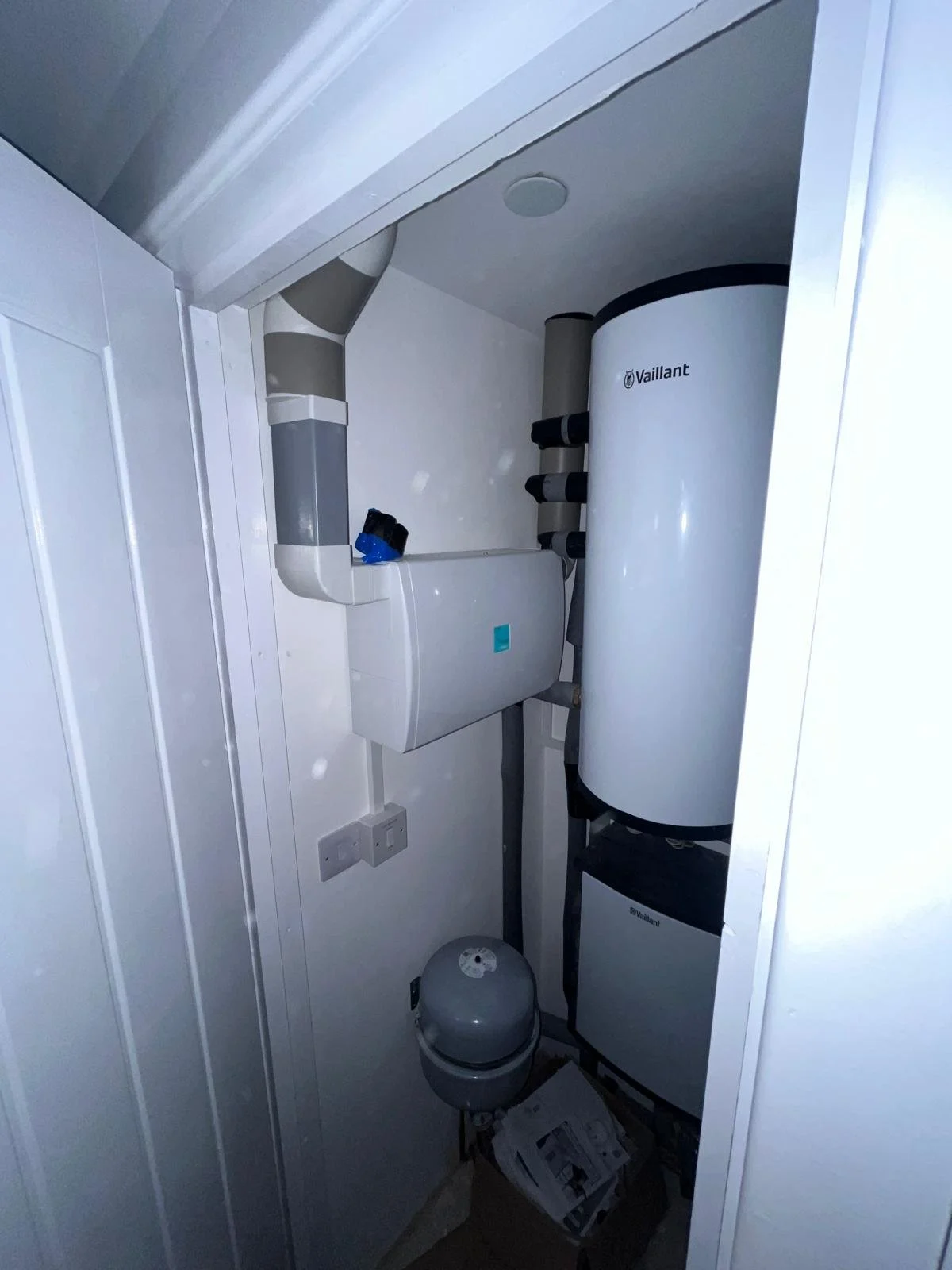 Utility closet with a Vaillant water heater, piping, and electrical outlets, small space with a wall-mounted box and a pressure tank on the floor.