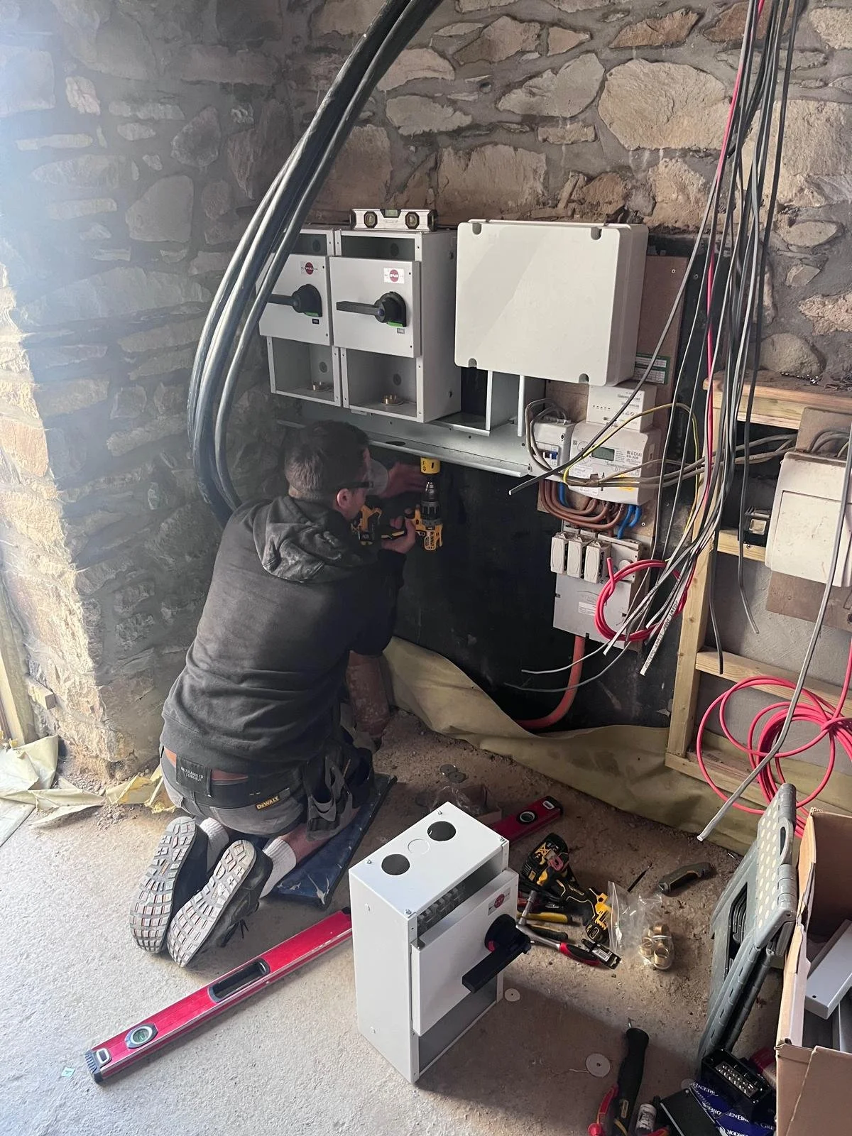 A person wearing safety glasses is working on electrical and plumbing components in a basement or utility room, kneeling on the floor with tools scattered around.