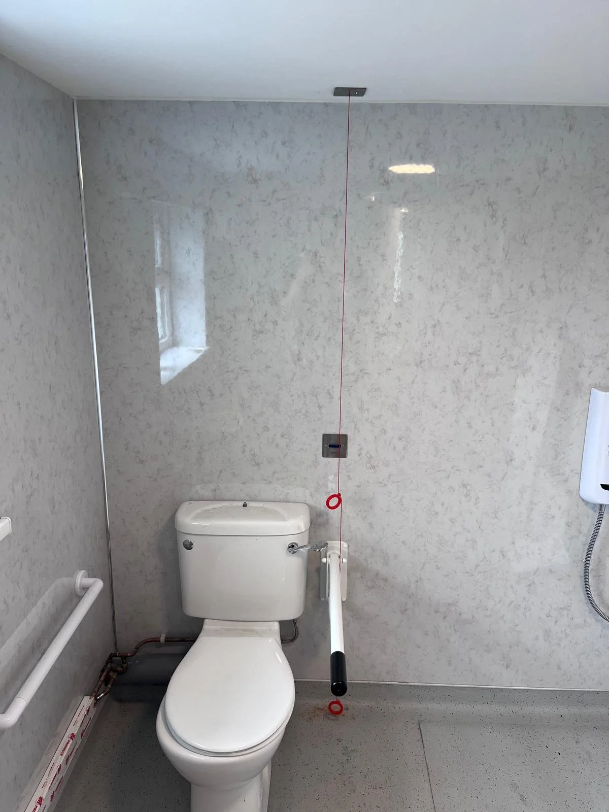 A hospital bathroom with a white toilet, a wall-mounted handrail, a small window on the left, and a wall-mounted hand sanitizer dispenser on the right. There is a red cord with a black handle hanging from the ceiling.