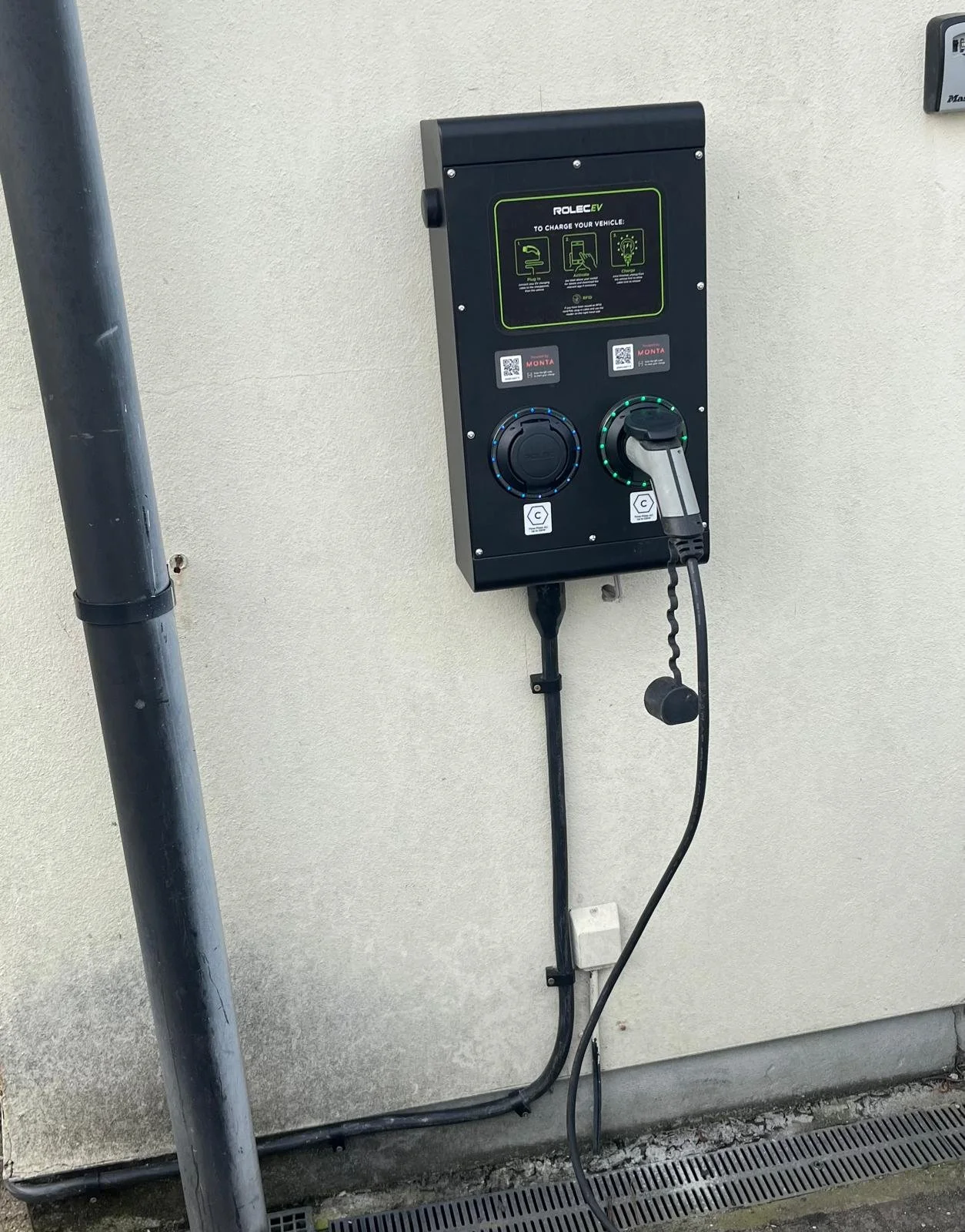 An electric vehicle charging station mounted on a beige exterior wall, with two charging connectors, one plugged in, and instructions on the front panel.