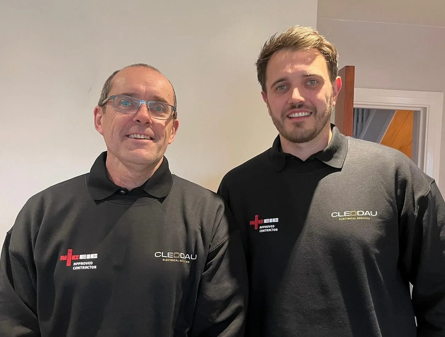 Two men standing side by side inside a building, both wearing black polo shirts with company logos. One man has glasses and short hair, the other has a beard and longer hair.