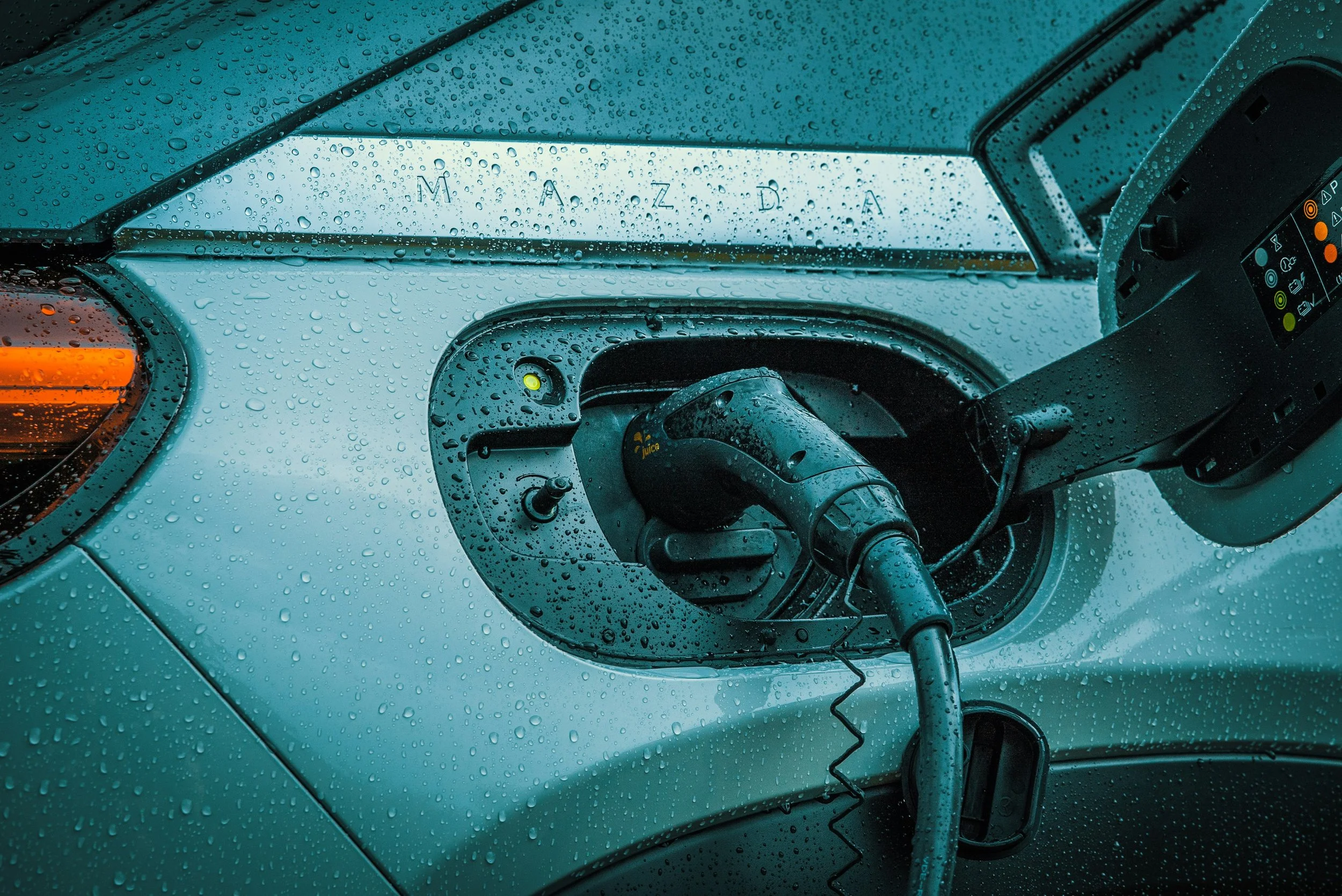 Electric vehicle parked outside on a rainy day, with rain droplets on the hood and charging port open, connected to a charging cable.