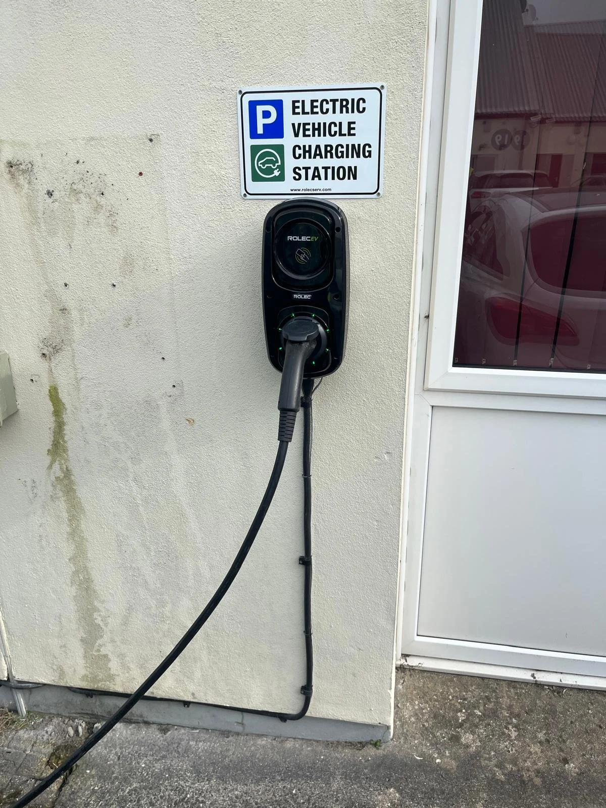 Electric vehicle charging station mounted on a wall next to a parking sign that reads 'Parking, Electric Vehicle Charging Station.' The station is black with a charging cable.