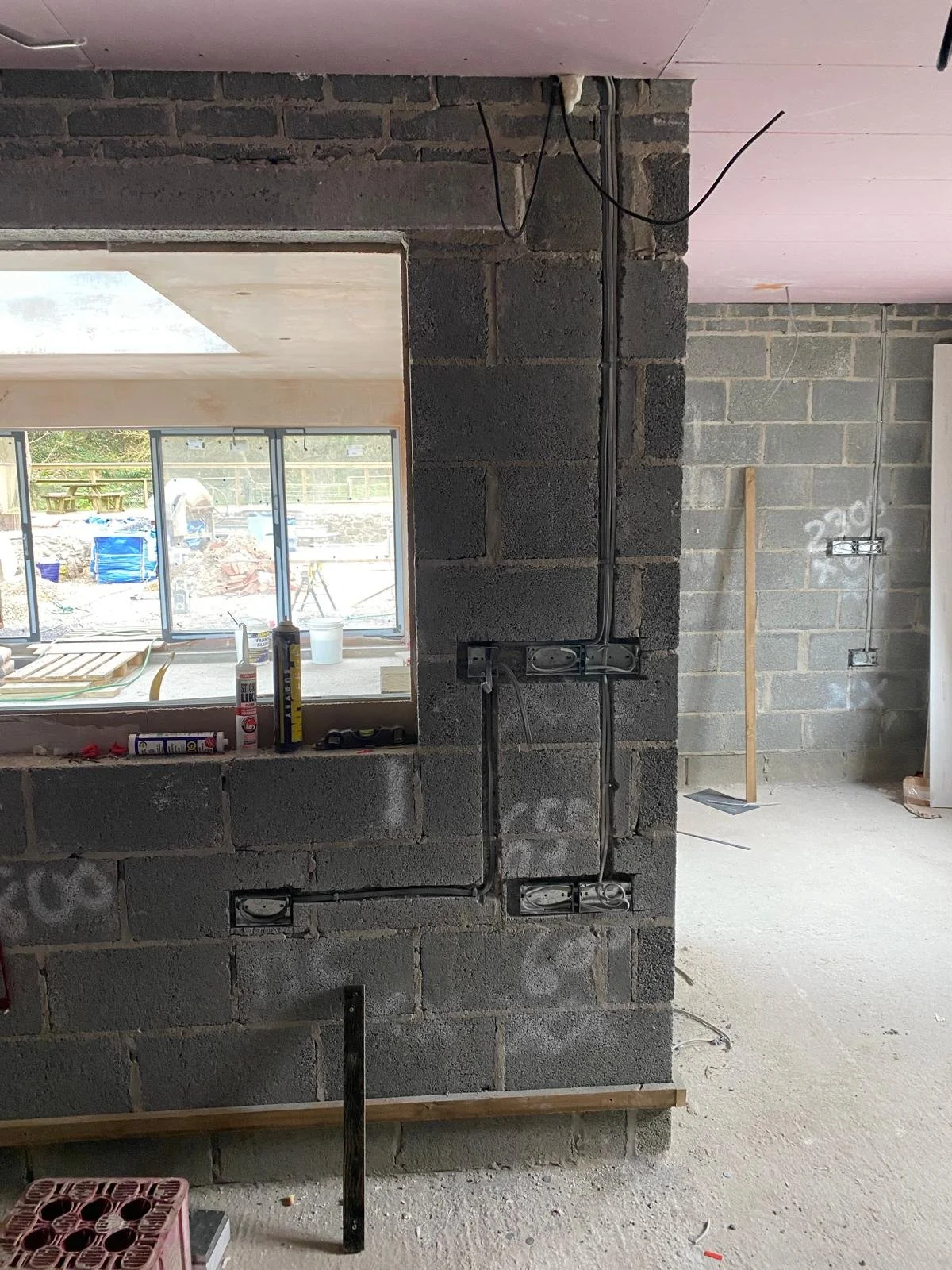 Interior of a house under construction with exposed brick walls, electrical wiring, and outlets being installed. Construction tools and supplies are on a ledge, and there is a large window with a view of an outdoor area with tools and building materials.