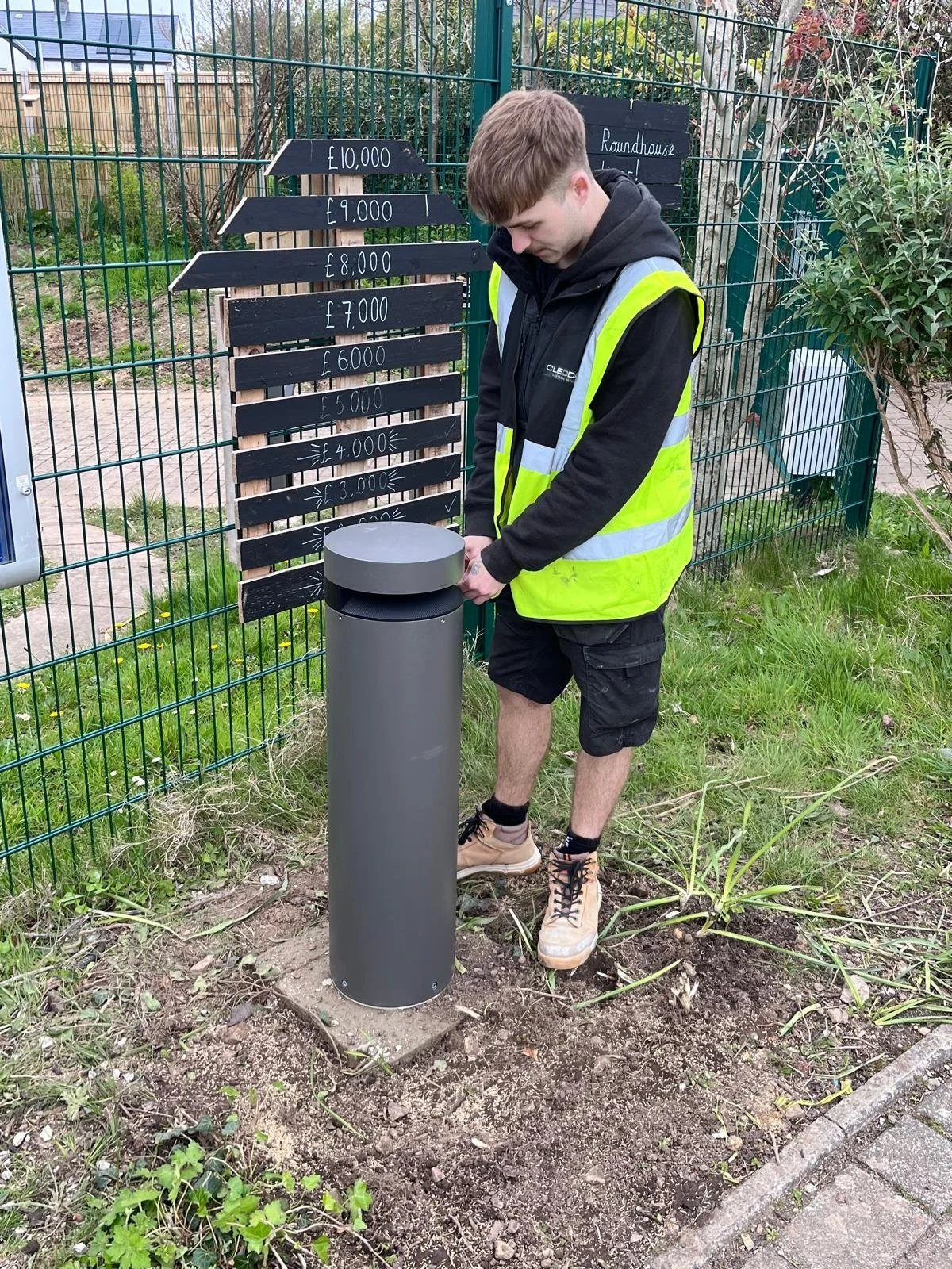 A person in a yellow reflective safety vest and black shorts is standing outdoors, adjusting a cylindrical trash or recycling bin. Behind him, there is a black directional sign with white lettering showing prices from £3,000 to £10,000 for different items, and a sign indicating a 'Roundhouse.' The area is enclosed with green fencing and has grass and plants on the ground.
