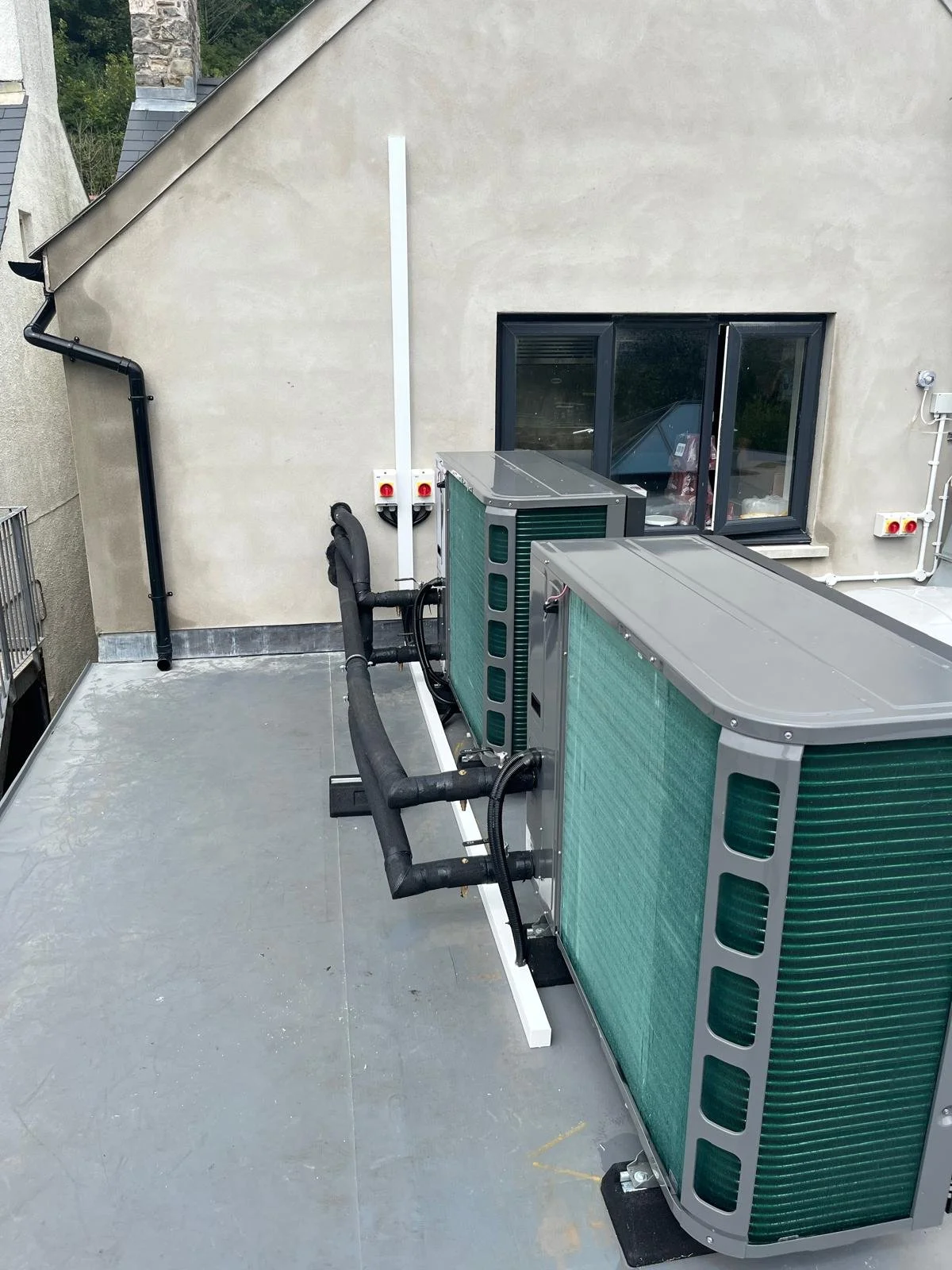 Rooftop with two large air conditioning units, black insulated pipes, and a wall with two electrical outlets and a window.