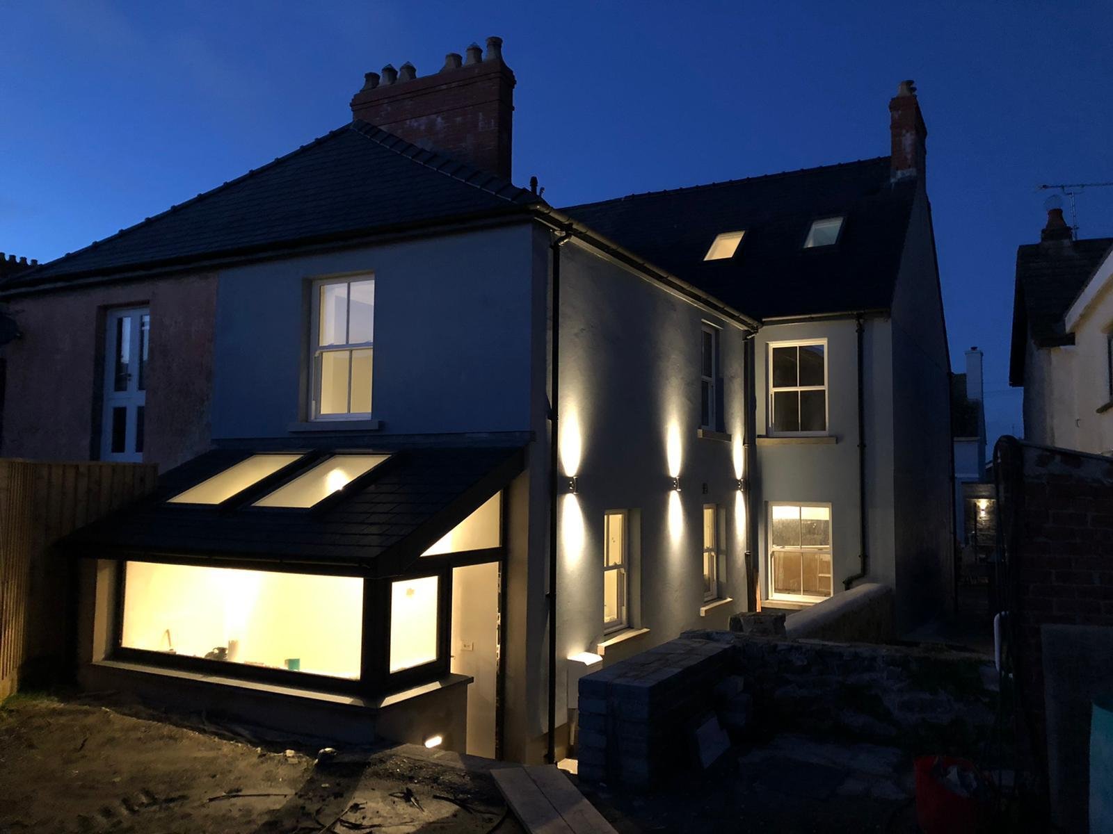 A multi-story house at dusk with exterior lights on, white walls, black roof, several windows, including a glass extension area, and chimney stacks, with neighboring houses visible.
