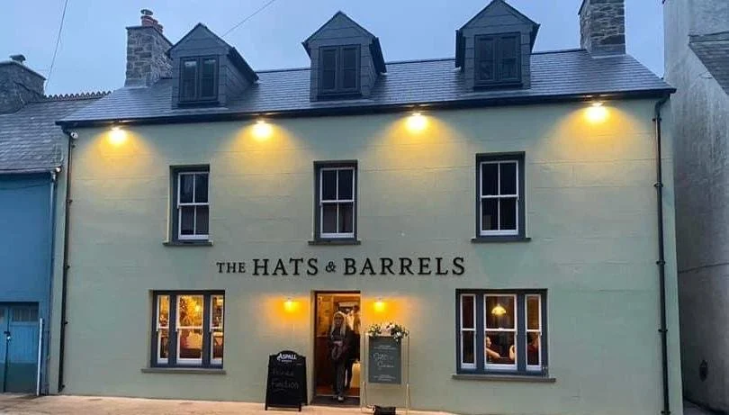 Front of a light green building with four upper windows, three lower windows, and a sign reading 'The Hats & Barrels', with a person standing at the entrance.