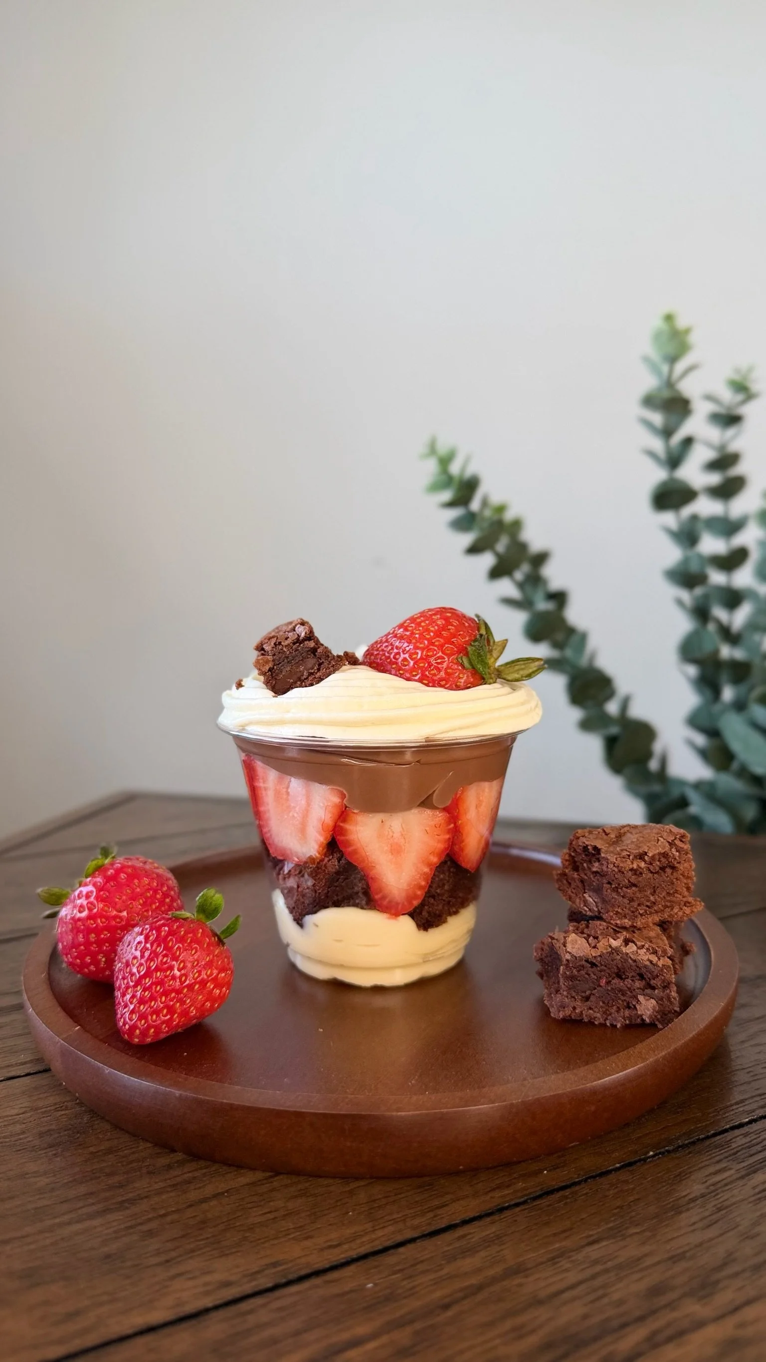 Strawberry parfait with layers of strawberries, chocolate brownie pieces, and whipped cream in a glass, topped with a whole strawberry and brownie chunks, surrounded by additional strawberries and brownie pieces on a wooden tray.