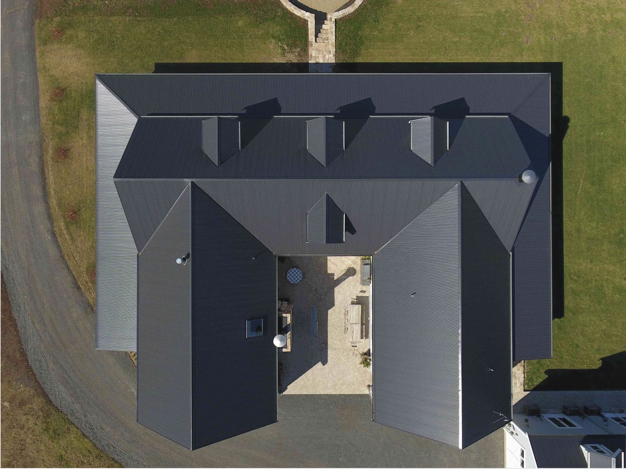 An aerial view of a large Whangamata house with a dark metal roof, surrounded by green lawn and a gravel driveway. The house has multiple gabled sections, small chimneys, and a patio area with outdoor furniture and a fire pit.