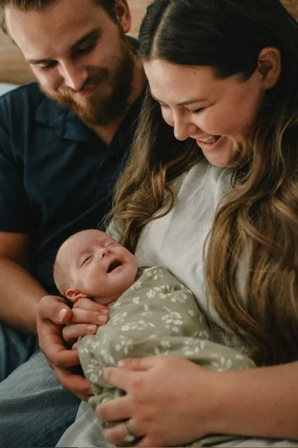A woman holding a baby while a man looks on happily.