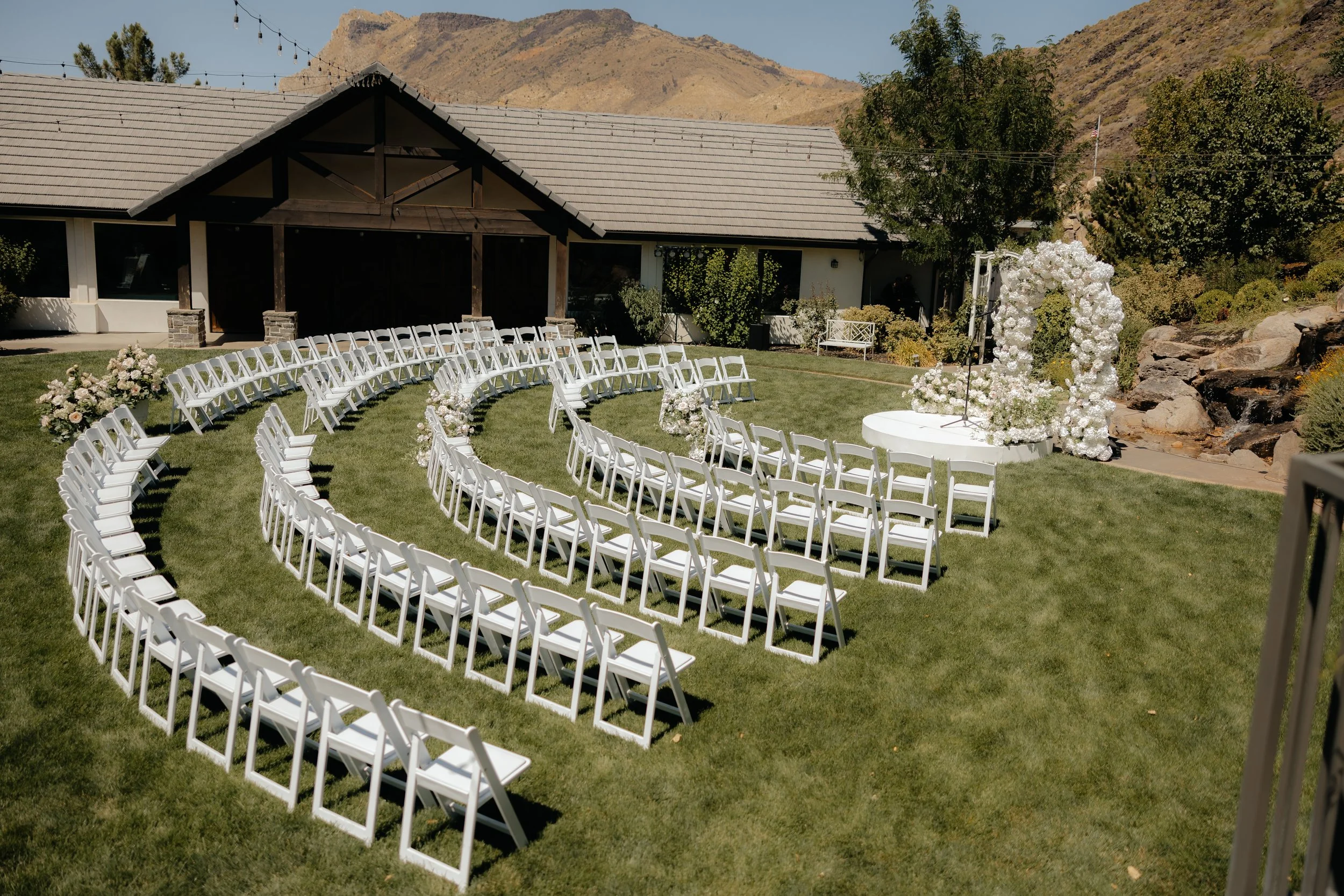 Outdoor wedding setup with white chairs arranged in a semi-circle on a grassy lawn, decorated with floral arrangements and a floral arch, with a mountain backdrop.