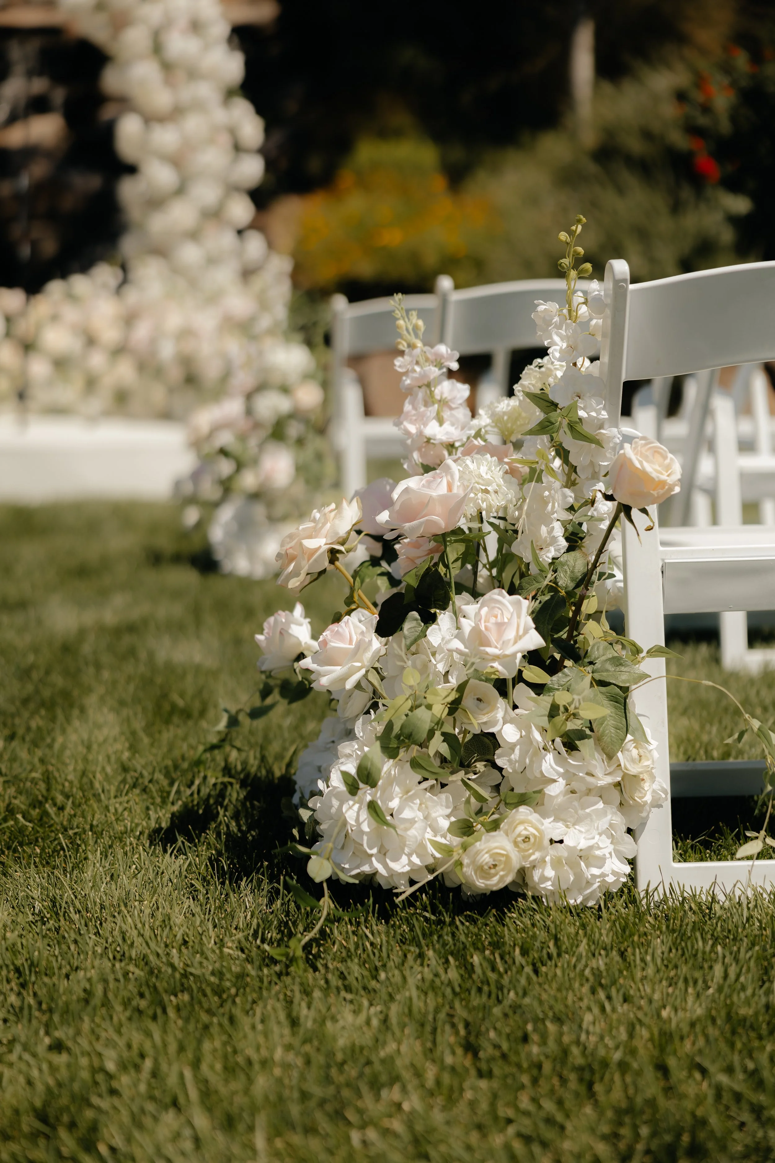 White chairs and a floral arrangement of white and light pink flowers at an outdoor wedding or event.