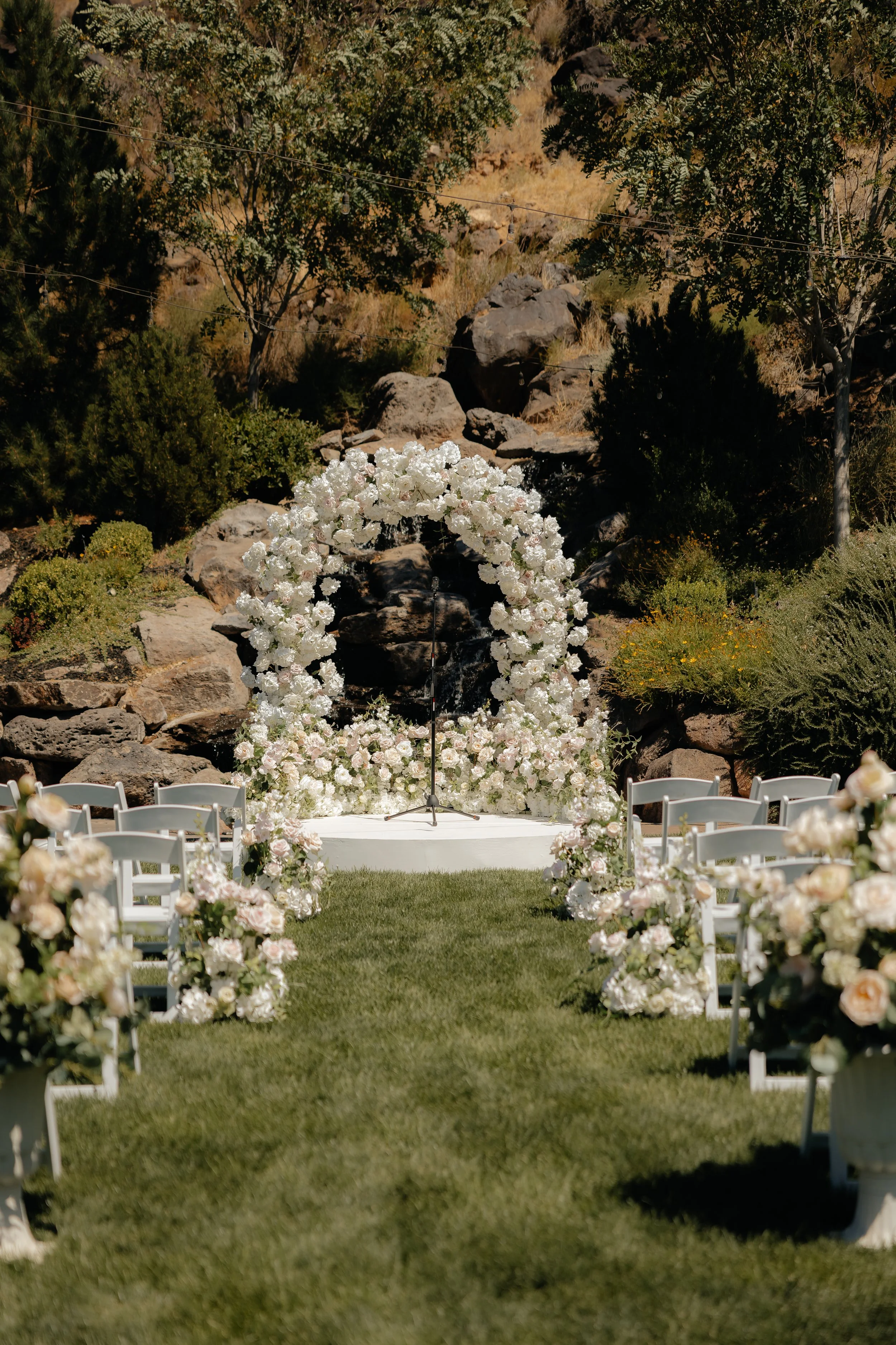 Outdoor wedding ceremony setup with white floral arch, white chairs, and floral arrangements, on a grassy aisle, with trees and rocks in the background.