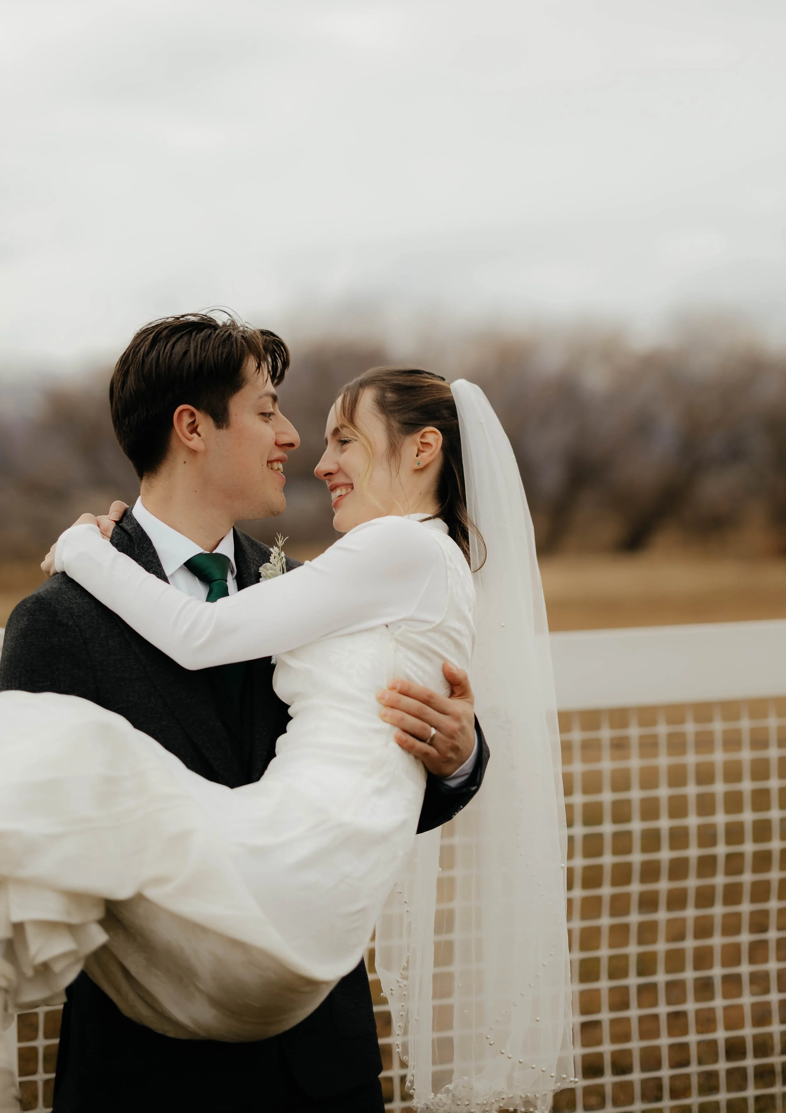 A groom lifting a bride, both smiling and looking at each other, outdoors on a cloudy day with trees and a white fence in the background.