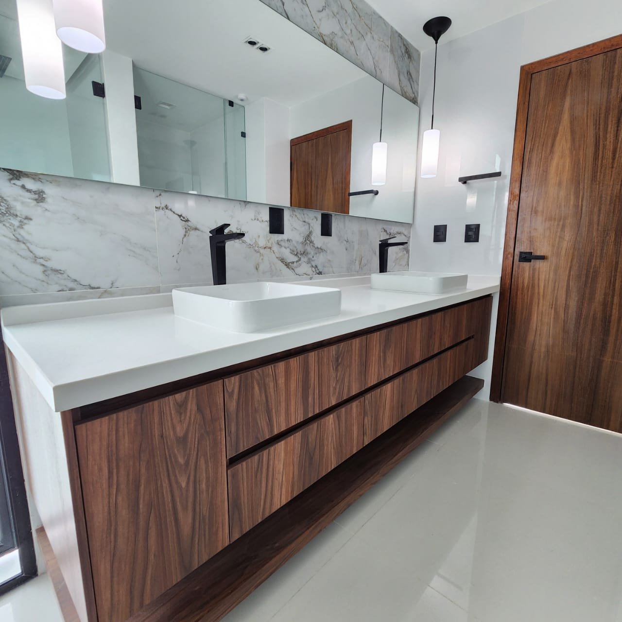 Lavabo doble con mesada de piedra blanca y gabinetes de madera, espejo grande, puertas de madera, luces colgantes y accesorios negros en un baño moderno.