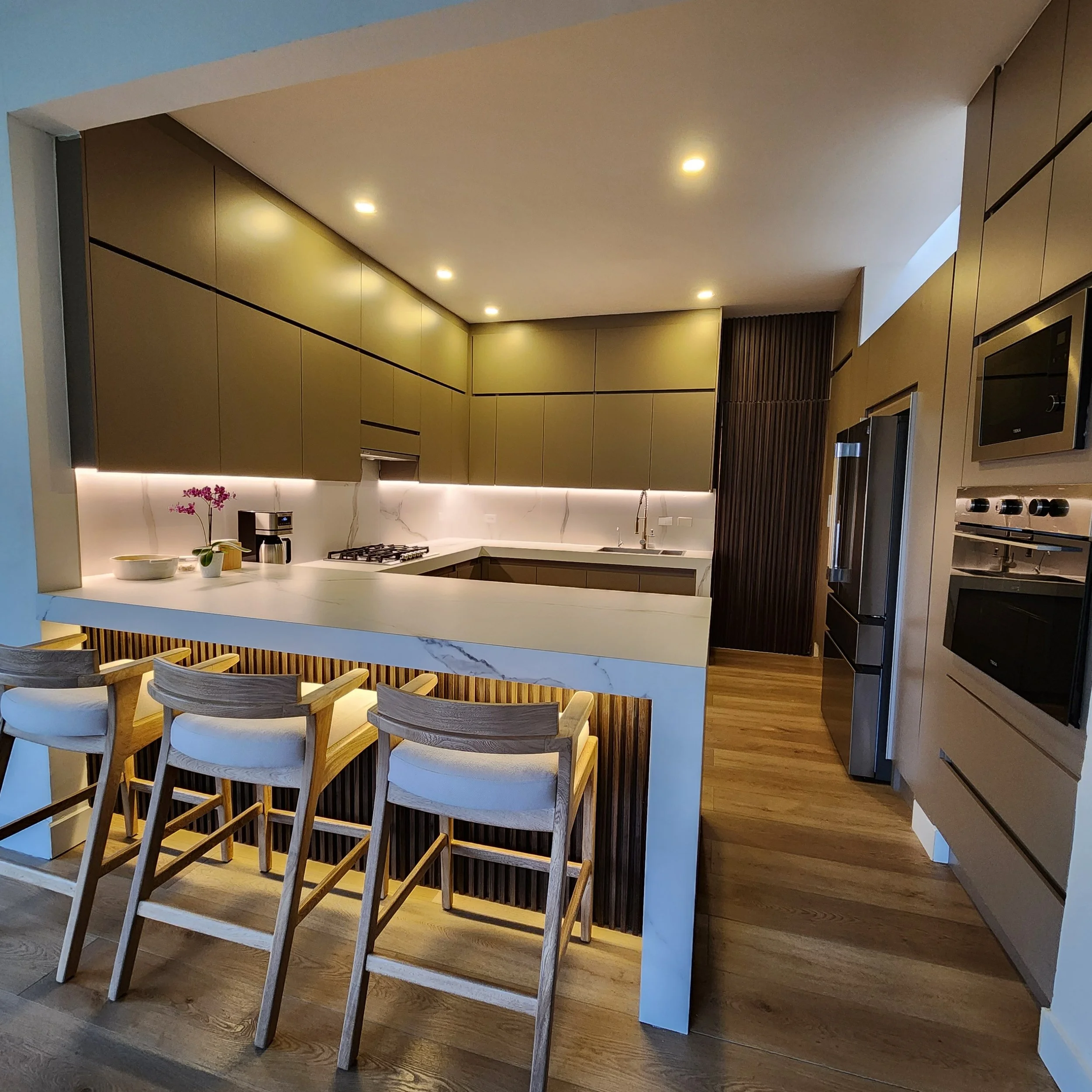 interior de una cocina moderna con gabinetes beige, mesón de mármol claro, y barra con sillas de madera claras. Hay una planta de orquídea en una taza blanca sobre la mesa y un electrodoméstico en la encimera.