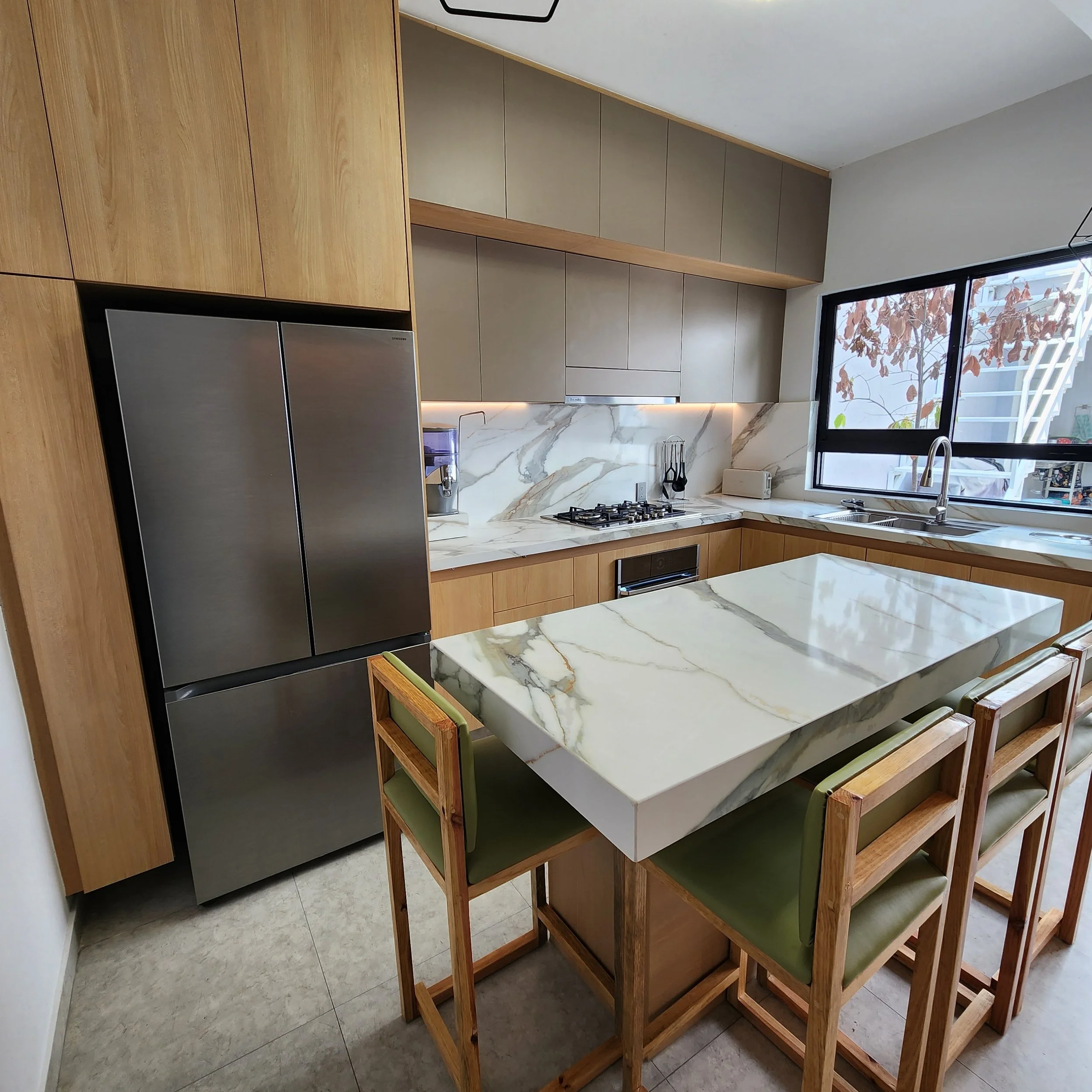 Cocina moderna con gabinetes de madera, superficie de encimera de mármol y una mesa de comedor de madera con sillas con cojines verdes, cerca de una ventana que muestra árboles con hojas marrones.
