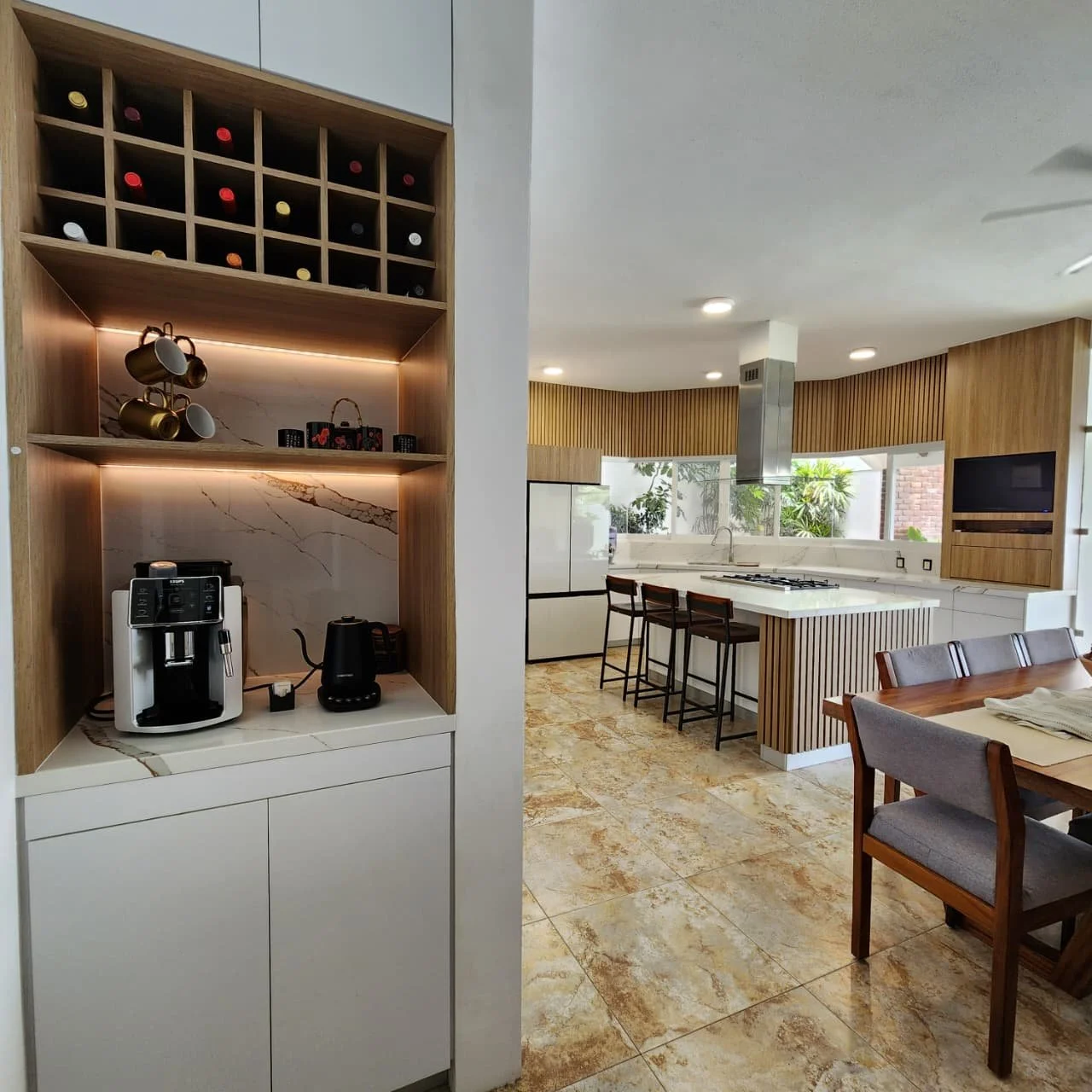 Cafetera y tetera en estantería en una cocina moderna con encimeras blancas, paredes de madera y sillones en la barra de la cocina.