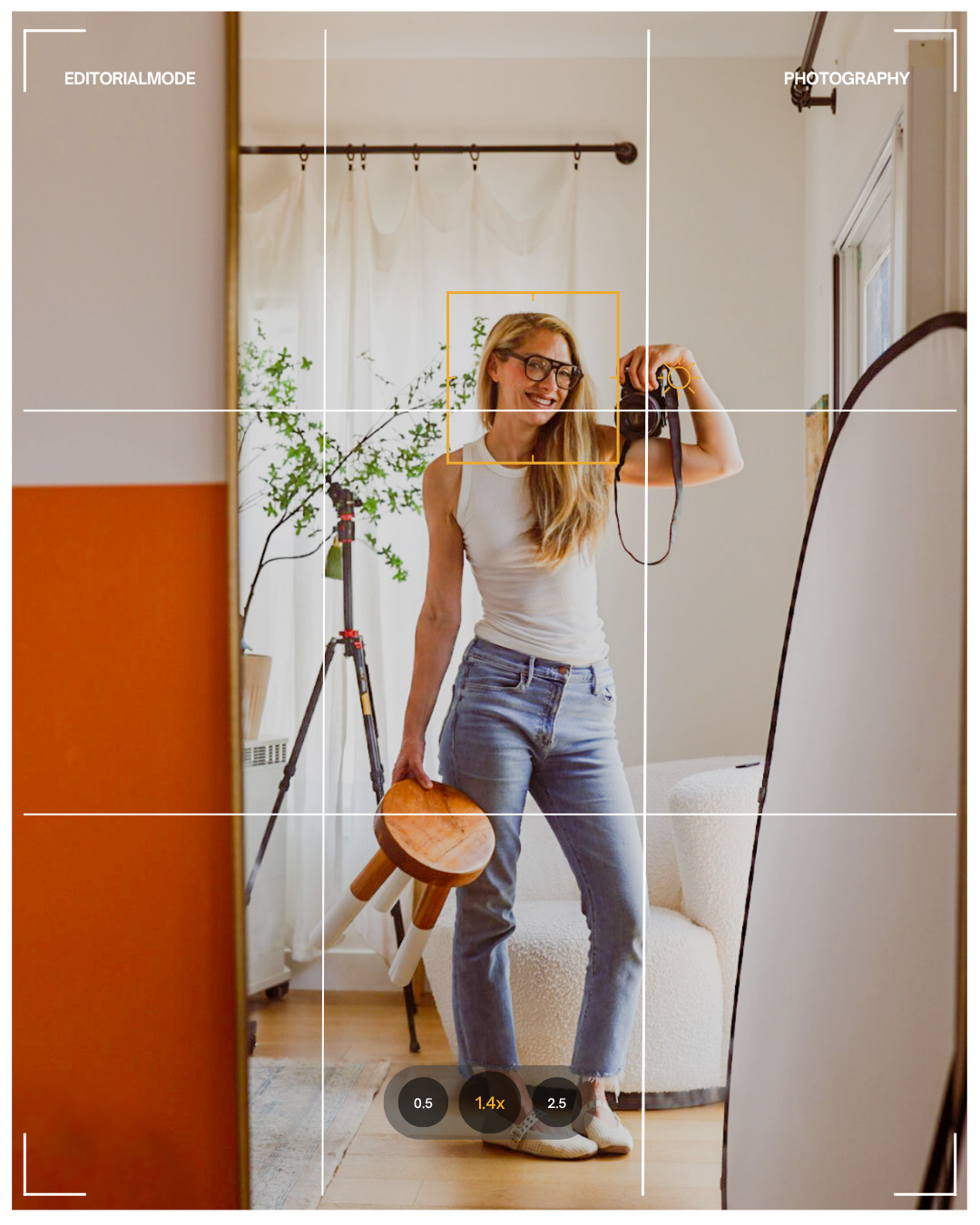 A woman with long blonde hair, glasses, wearing a white tank top and jeans, standing in front of a mirror, smiling, holding a camera in her right hand and a small round wooden stool in her left hand, inside a well-lit room with a plant and a window i