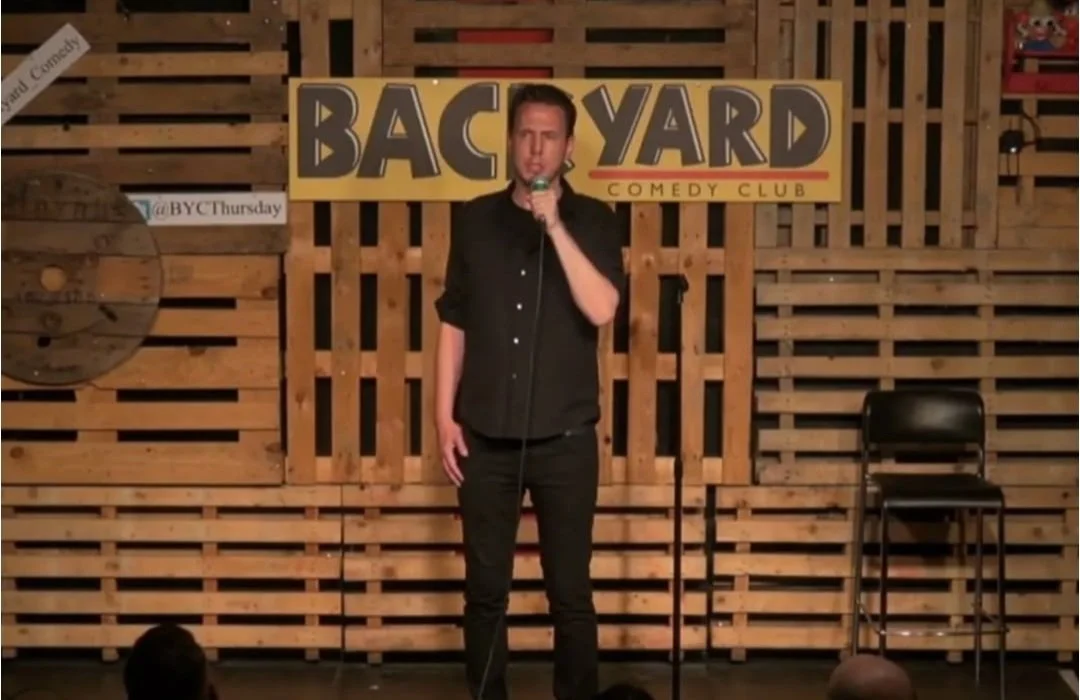 Male comedian standing on stage with a microphone at Backyard Comedy Club, with a wooden backdrop and a sign behind him.