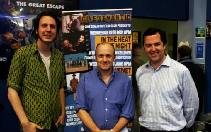Three men standing in front of a poster for a film club event at a cinema, smiling for the photo.