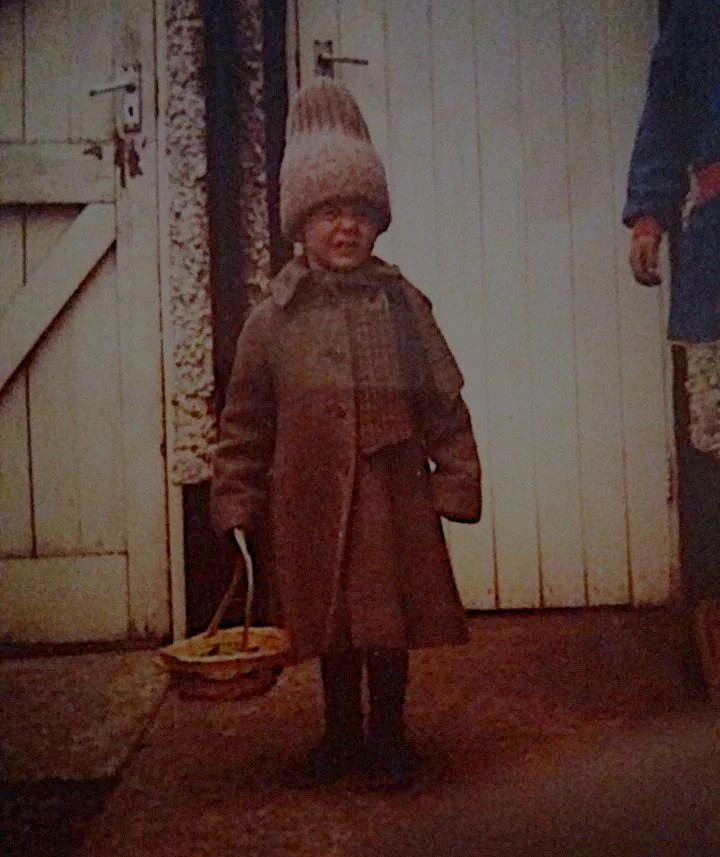 A young child with a distressed expression, wearing a brown coat, a knit hat, and holding a small basket indoors.