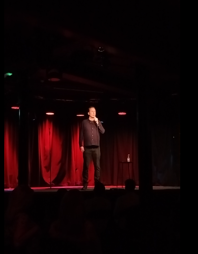 A comedian performing on stage with a red curtain backdrop, holding a microphone, with an audience in the foreground.