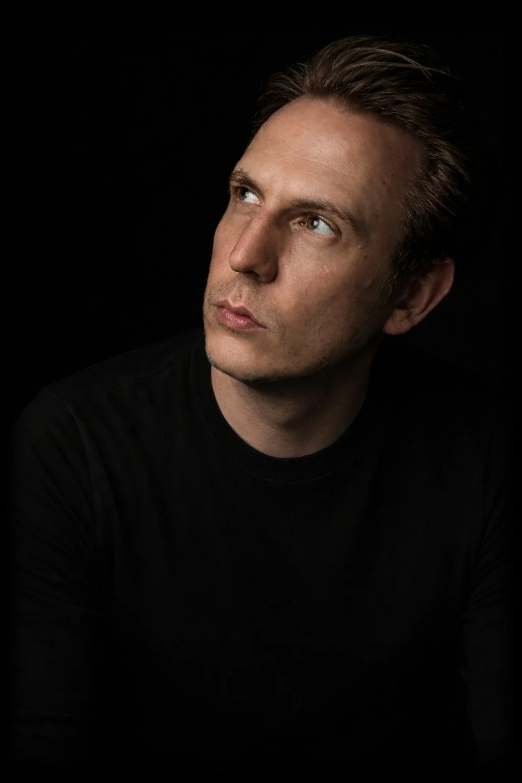 A man with light skin, brown hair, and blue eyes looking to his left, wearing a black shirt against a black background.