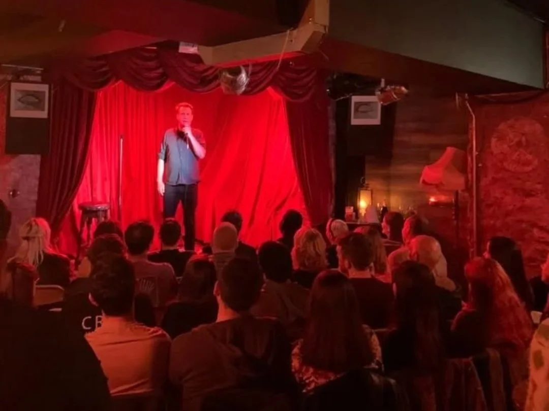 A comedian performs on stage in a dimly lit comedy club with red curtains, a microphone in hand, and an audience watching.
