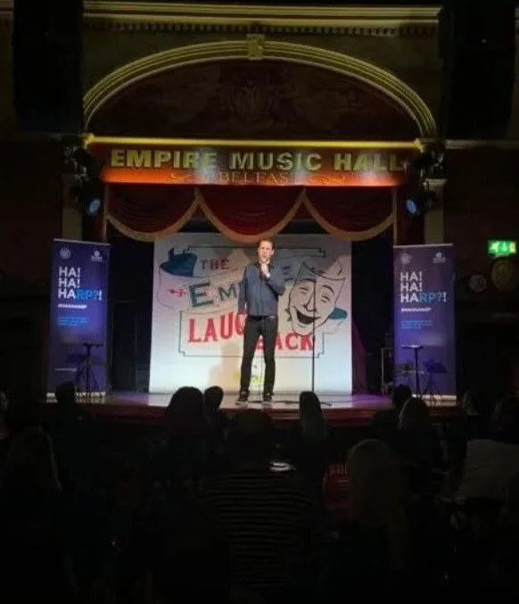 Stand-up comedian performing on stage at Empire Music Hall during The Laugh Attack event, with an audience watching.