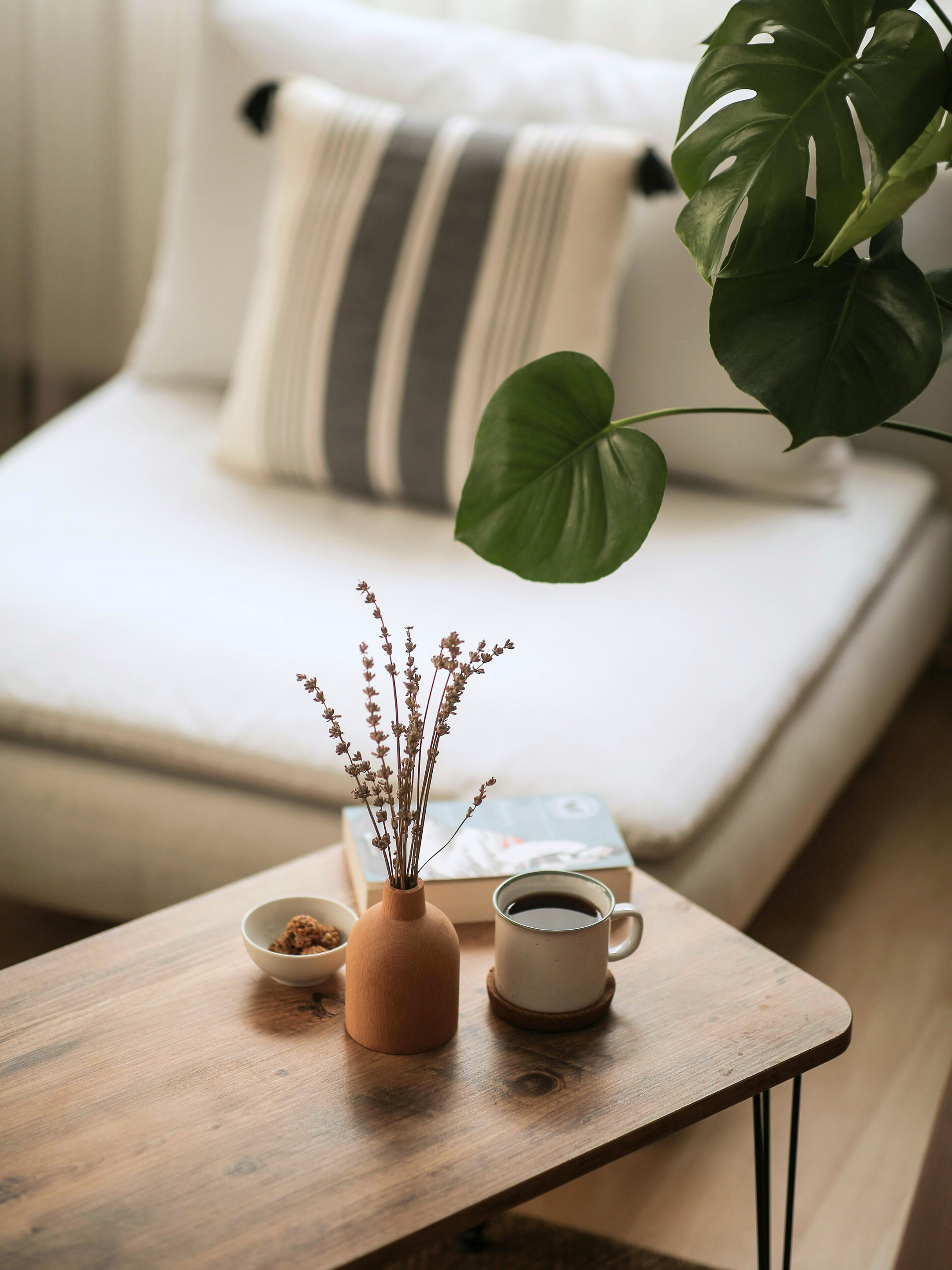 A cozy bedroom corner with a white bed and striped pillow, a wooden side table holding a green vase with dried flowers, a cup of coffee on a coaster, a small bowl, and a book. A large green leafy plant is in the foreground.