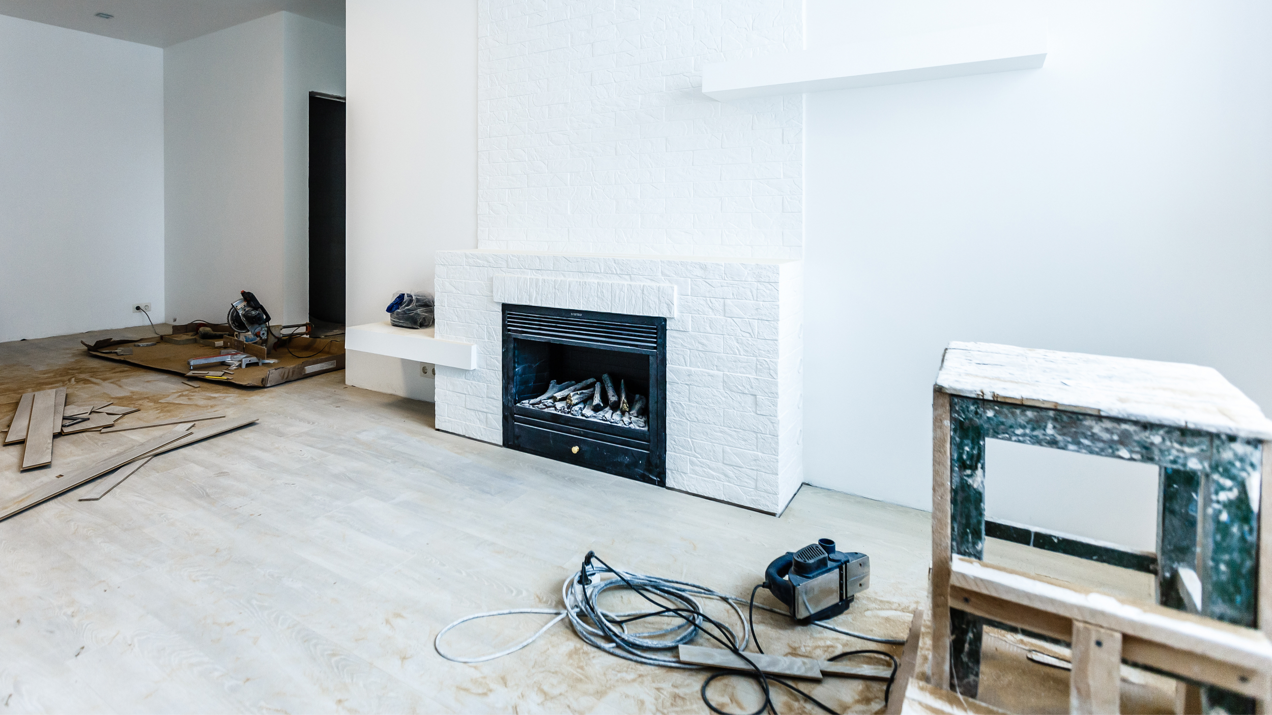 Living room under renovation with a white brick fireplace, construction tools, and unfinished flooring.
