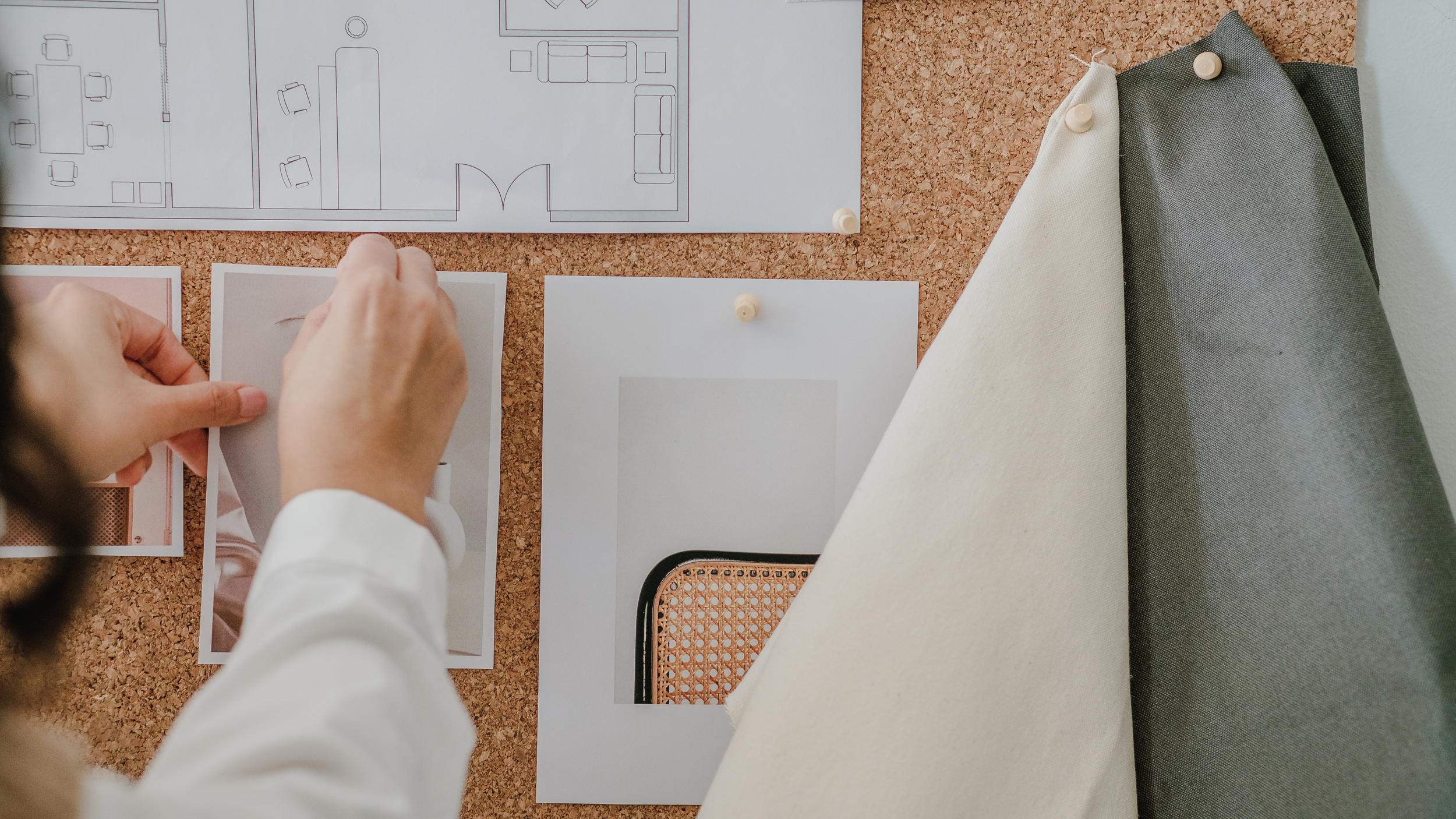 Person pinning a photograph on a corkboard with architecture plans and other photographs and fabric samples pinned nearby.