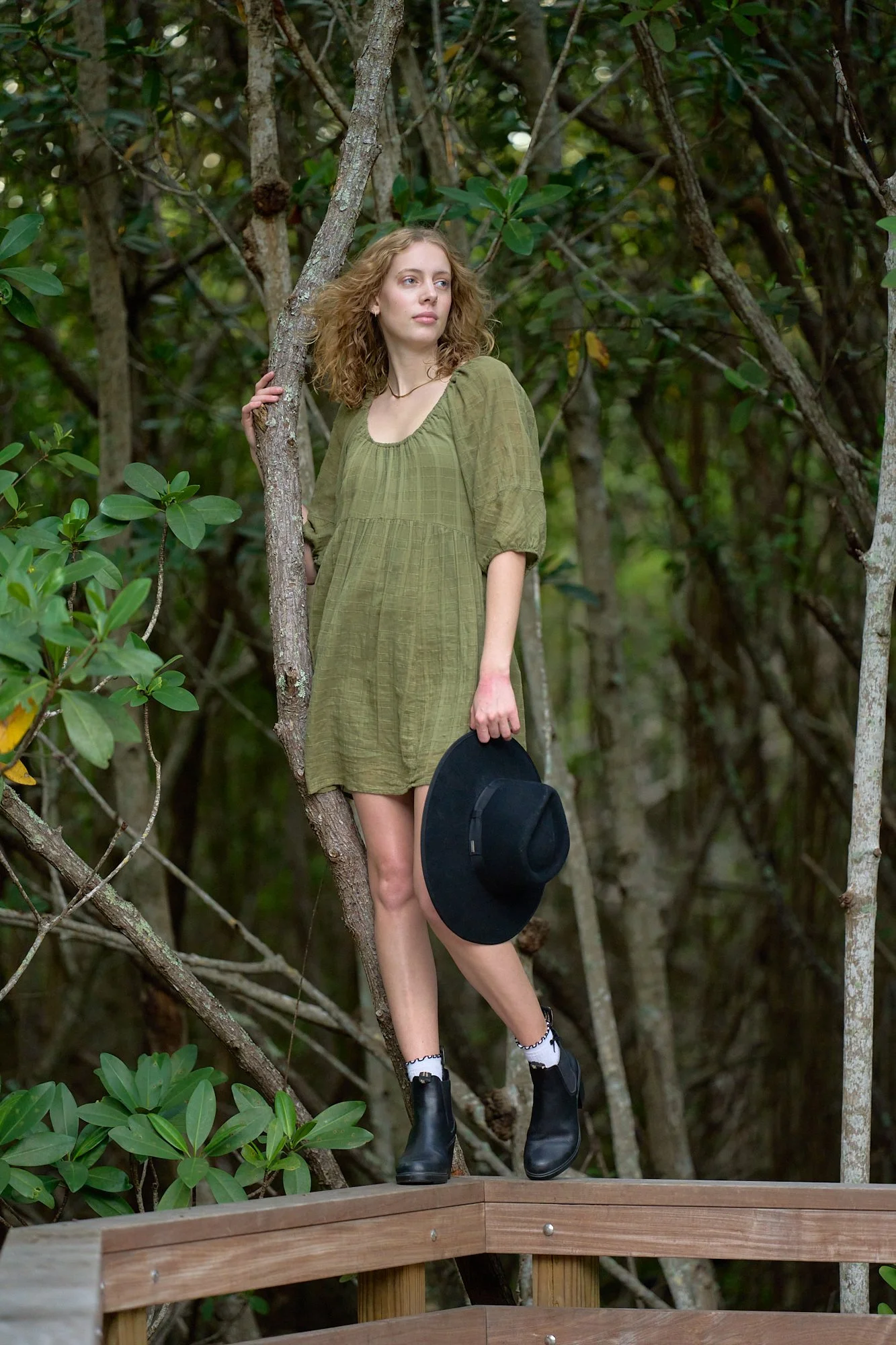 A young woman with curly blonde hair wearing a green dress, black ankle boots, and white socks stands on a wooden platform in a forest, holding a black hat and resting her hand on a tree branch.
