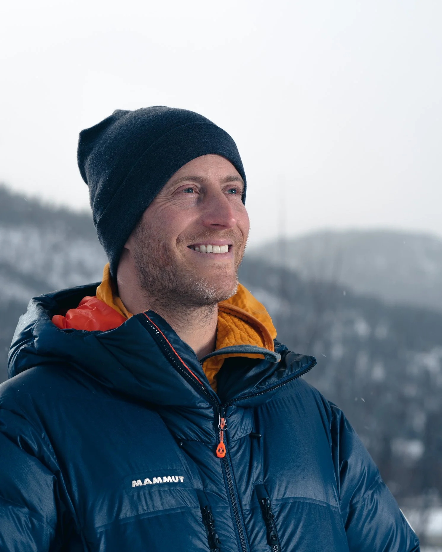 A man smiling outdoors in winter, wearing a black beanie, a navy blue puffy jacket, and an orange inner layer, with snow-covered hills in the background.
