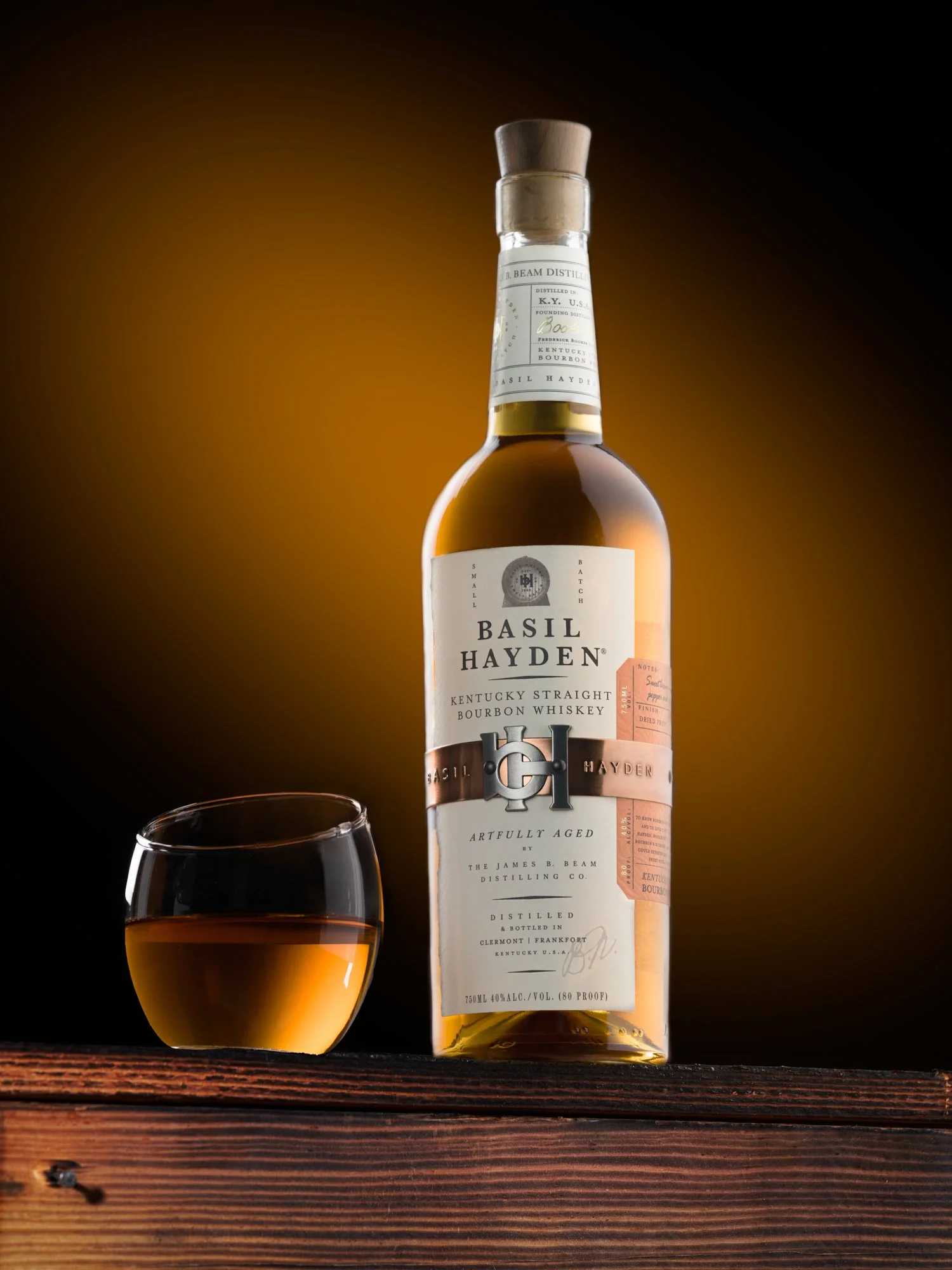 A bottle of Basil Hayden Bourbon Whiskey and a glass of whiskey on a wooden surface with a dark background and warm lighting.
