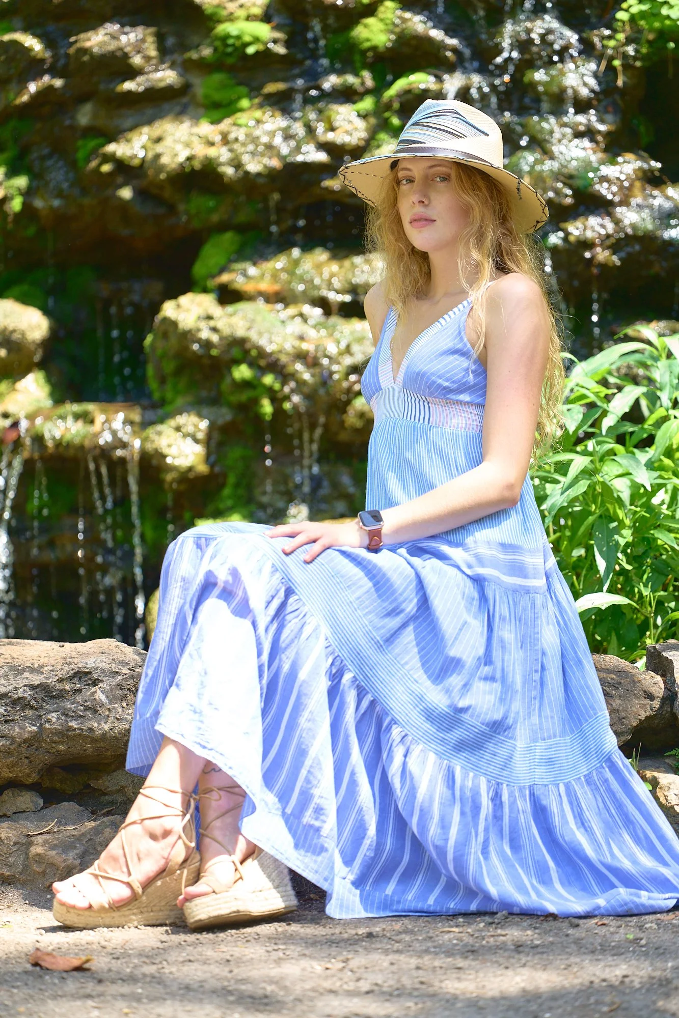 A young woman with long blonde hair wearing a wide-brimmed hat, a blue and white striped dress, and sandals, sitting on a rock beside a small waterfall in a lush outdoor setting.