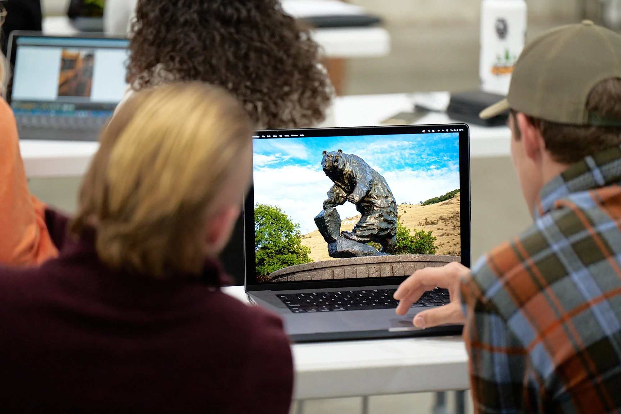People looking at a laptop displaying a statue of a bear on a pedestal in an outdoor setting.