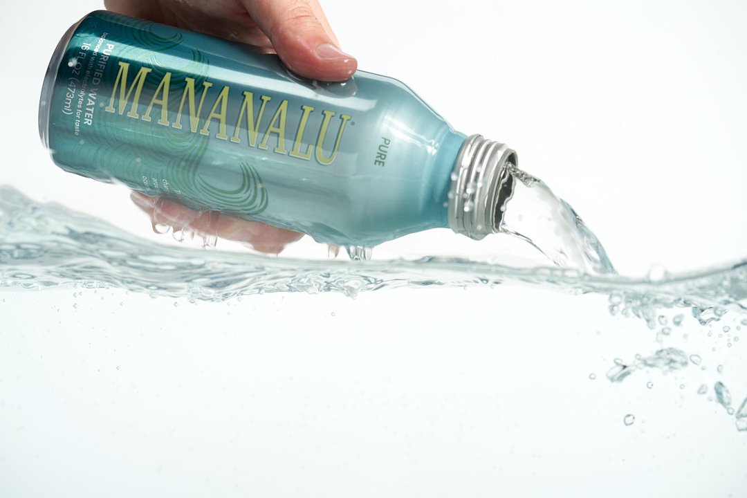 A hand holding a can labeled 'Mananalu' pouring water into a clear surface, with water splashing.